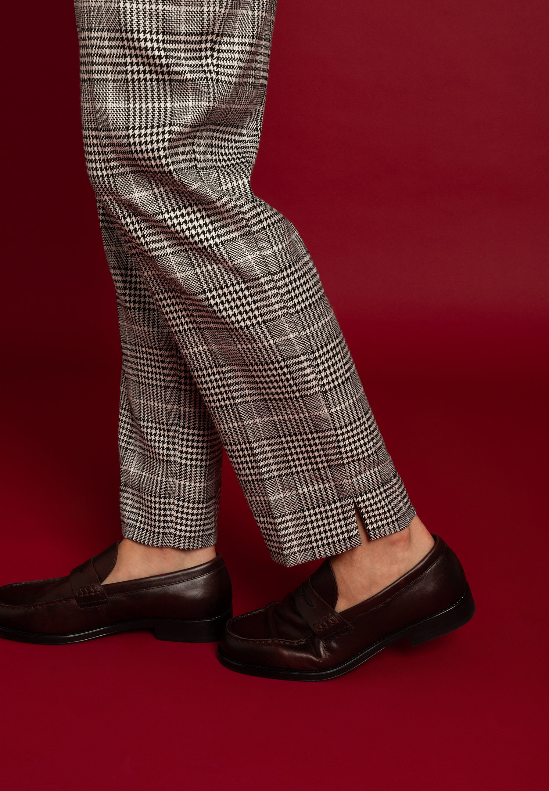 Nahaufnahme von MORE & MORE Hose mit Hahnentrittmuster und braunen Loafers auf rotem Grund. Nahaufnahme von MORE & MORE Hose mit Hahnentrittmuster und braunen Loafers auf rotem Grund.