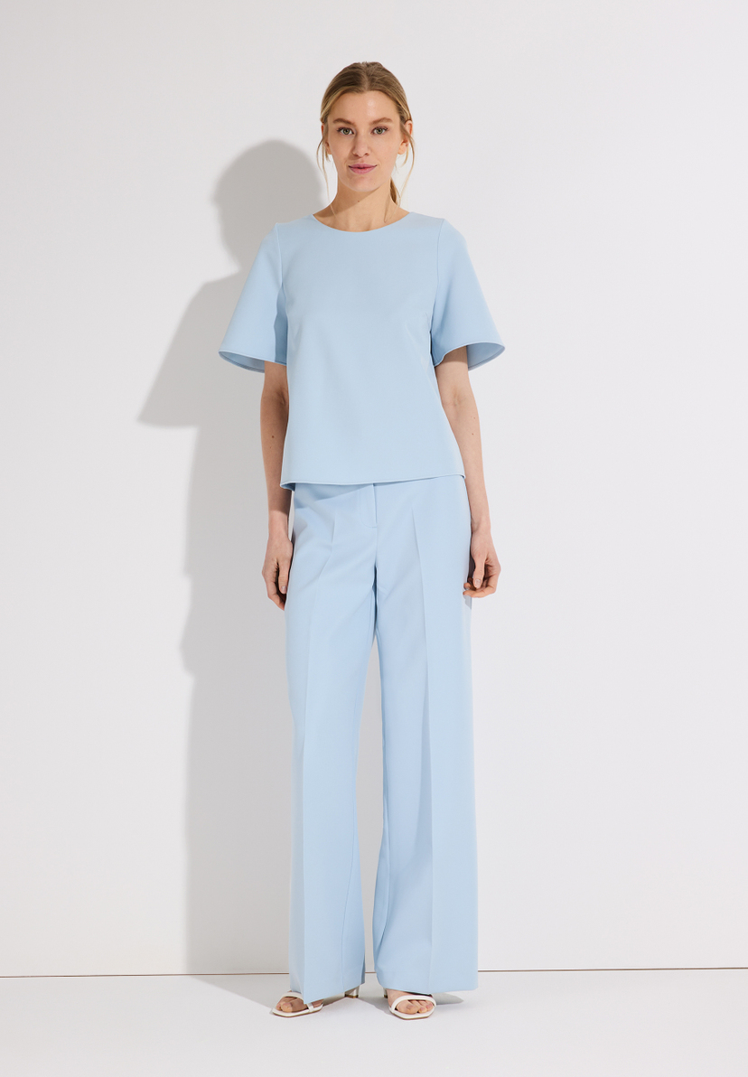 Eine Frau in einem hellblauen MORE & MORE Hosenanzug posiert vor einer weißen Wand. Eine Frau in einem hellblauen MORE & MORE Hosenanzug posiert vor einer weißen Wand.