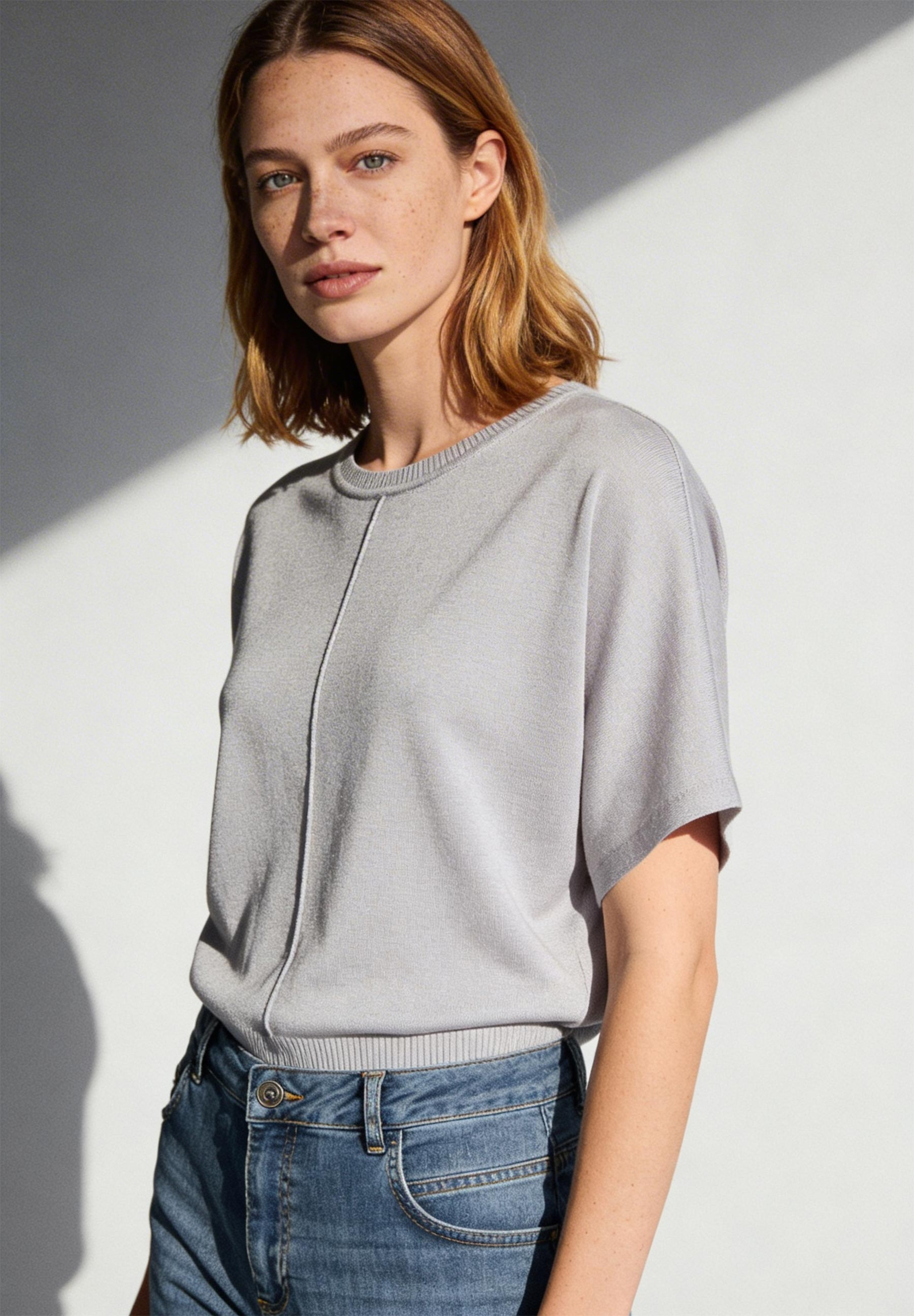 Eine Frau mit Sommersprossen trägt ein MORE & MORE Strickoberteil und Jeans vor einer weißen Wand.