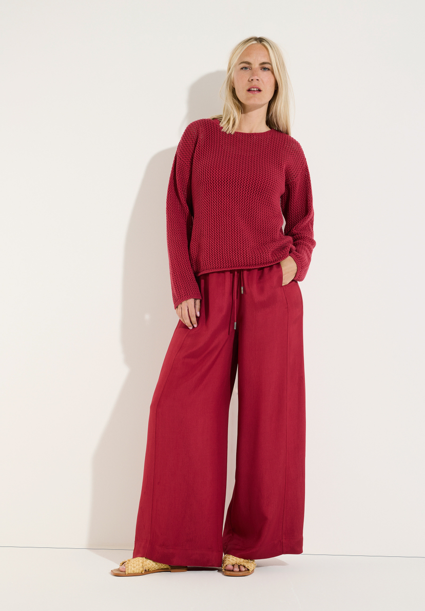 MORE & MORE Jeans: Blonde Frau in weinrotem Strickpullover u. passender weiter Hose posiert vor weißem Hintergrund. MORE & MORE Jeans: Blonde Frau in weinrotem Strickpullover u. passender weiter Hose posiert vor weißem Hintergrund.