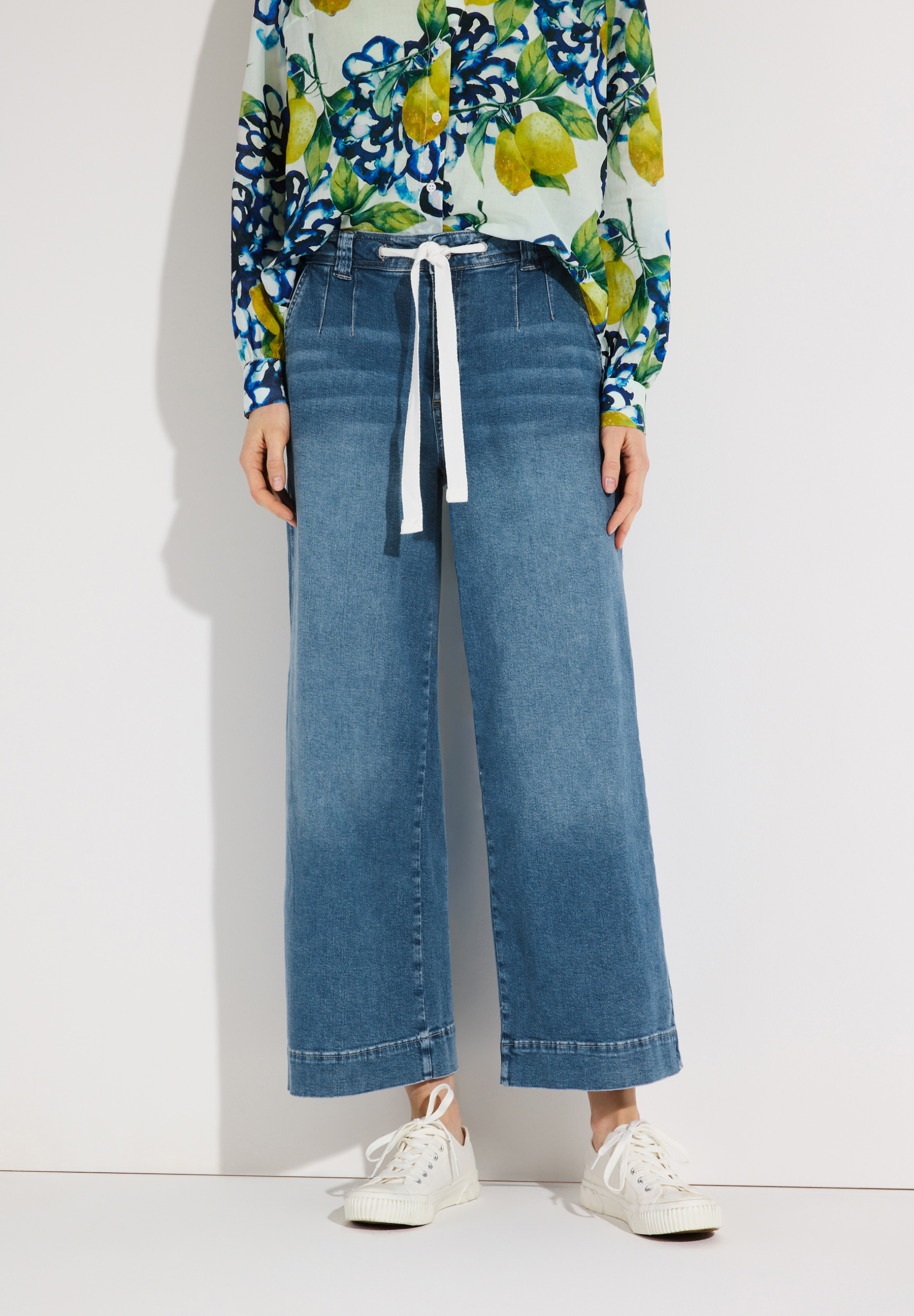 Eine MORE & MORE Jeans mit weitem Bein und weißem Kordelzug. Dazu eine Bluse mit Zitronenmuster und weiße Sneakers.