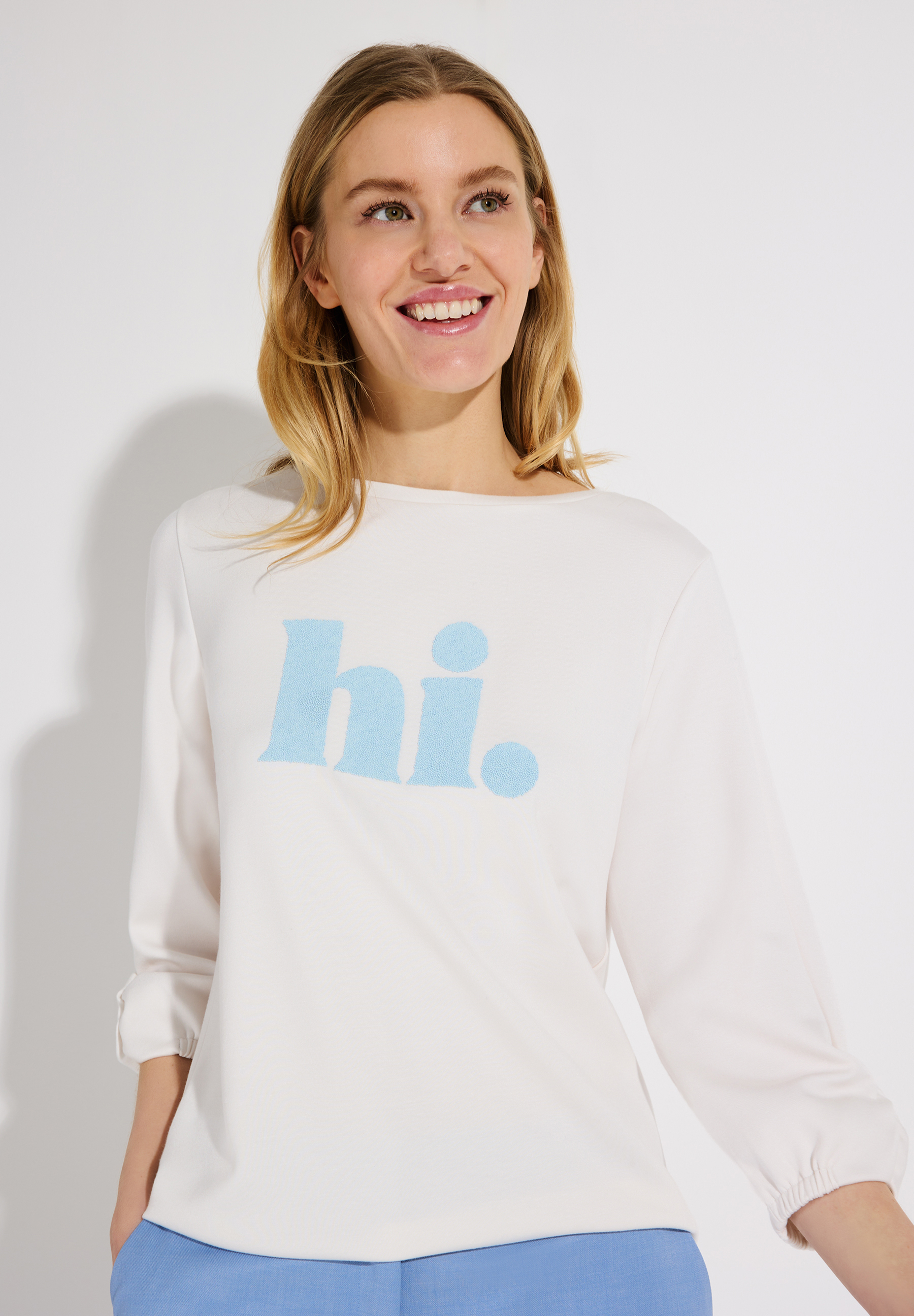 Eine lächelnde Frau trägt ein weißes MORE & MORE Sweatshirt mit dem blauen Aufdruck "hi."