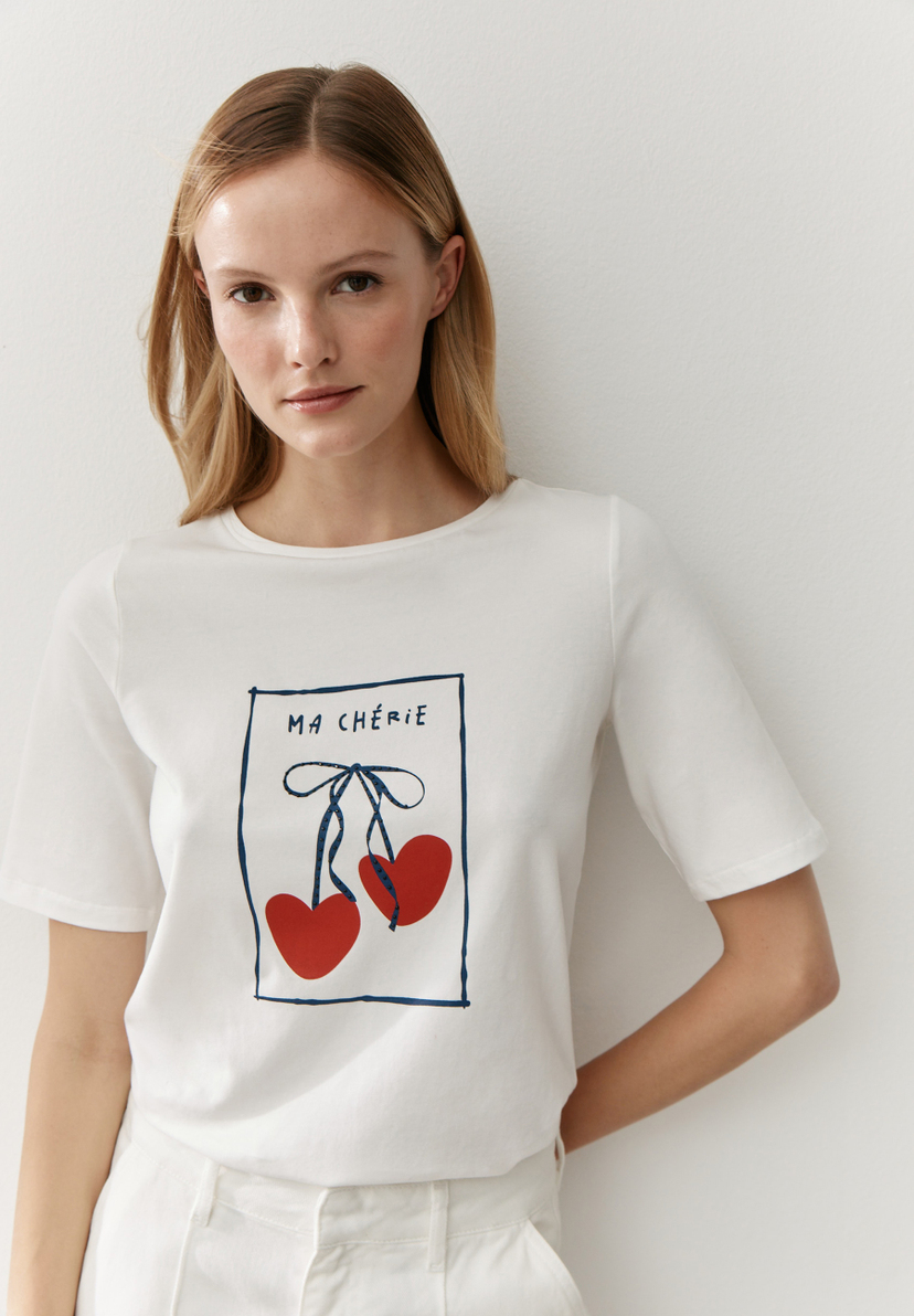 Eine Frau trägt ein weißes T-Shirt von MORE & MORE mit einem Kirschmotiv und dem Text "Ma Chérie". Eine Frau trägt ein weißes T-Shirt von MORE & MORE mit einem Kirschmotiv und dem Text "Ma Chérie".