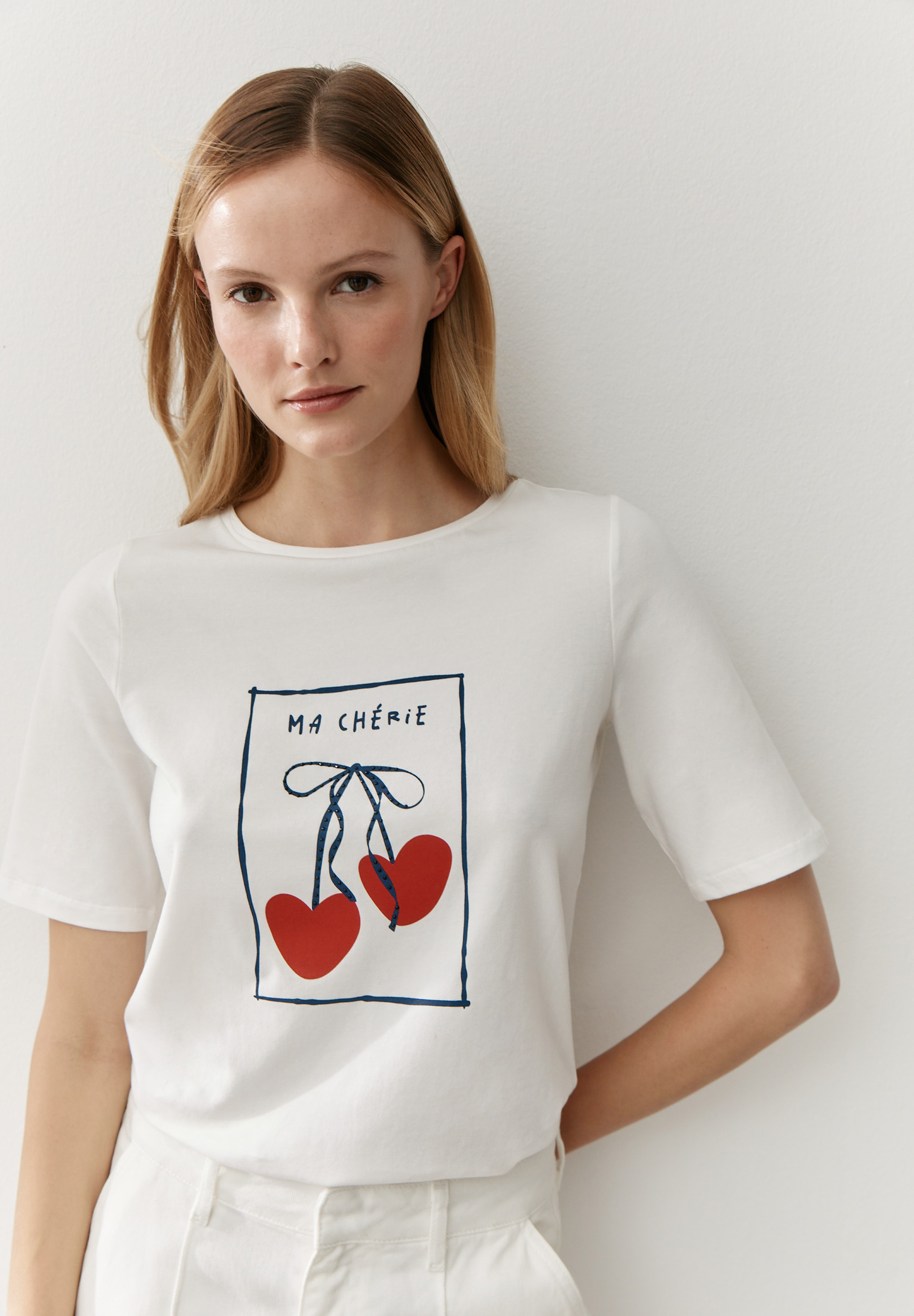 Eine Frau trägt ein weißes T-Shirt von MORE & MORE mit einem Kirschmotiv und dem Text "Ma Chérie".