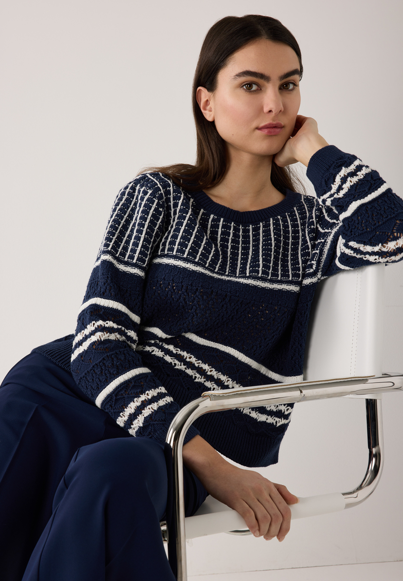 Eine Frau mit einem MORE & MORE Strickpullover sitzt auf einem Stuhl vor einem hellen Hintergrund. Eine Frau mit einem MORE & MORE Strickpullover sitzt auf einem Stuhl vor einem hellen Hintergrund.