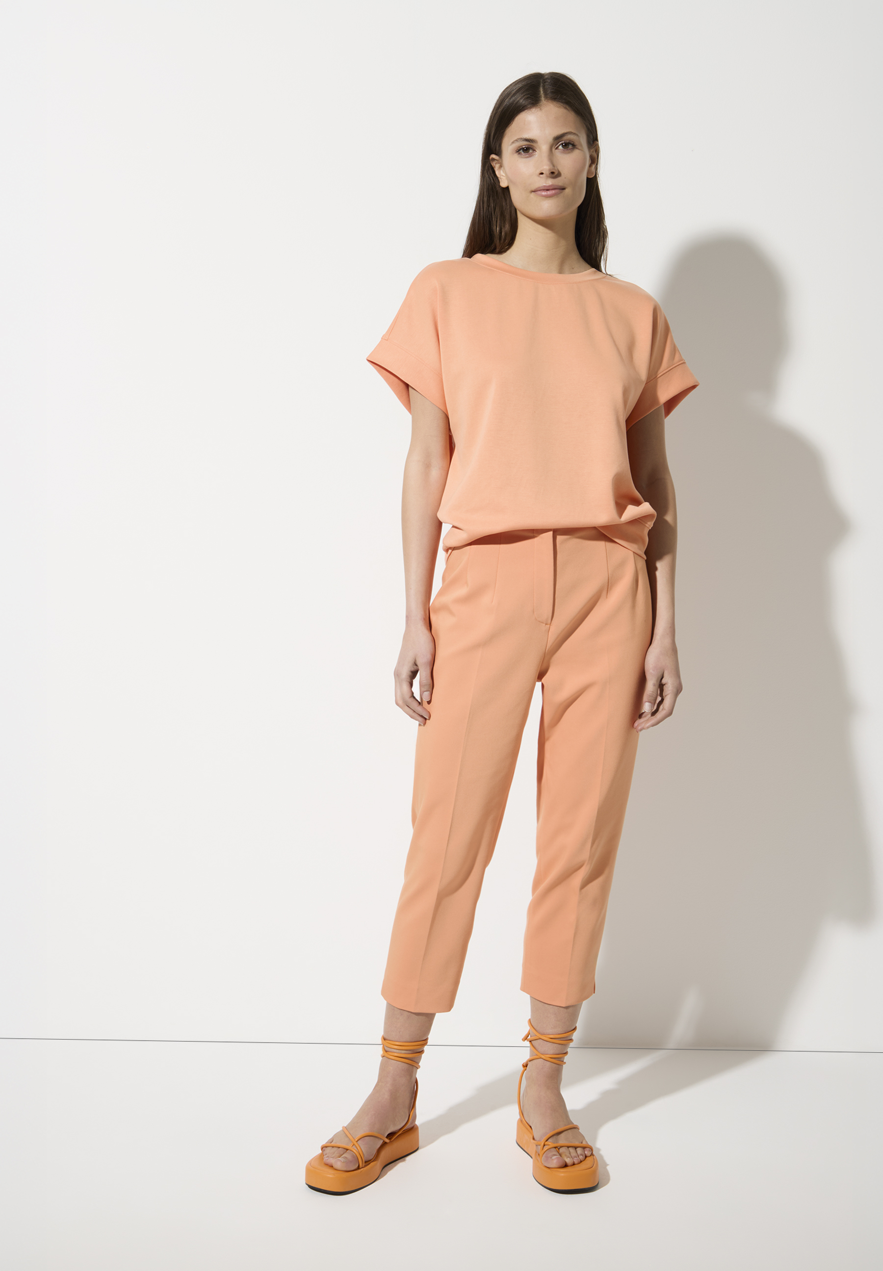 Eine Frau trägt ein orangefarbenes Hosen-T-Shirt-Set von MORE & MORE mit Baumwollmischung und passende Sandalen. Eine Frau trägt ein orangefarbenes Hosen-T-Shirt-Set von MORE & MORE mit Baumwollmischung und passende Sandalen.