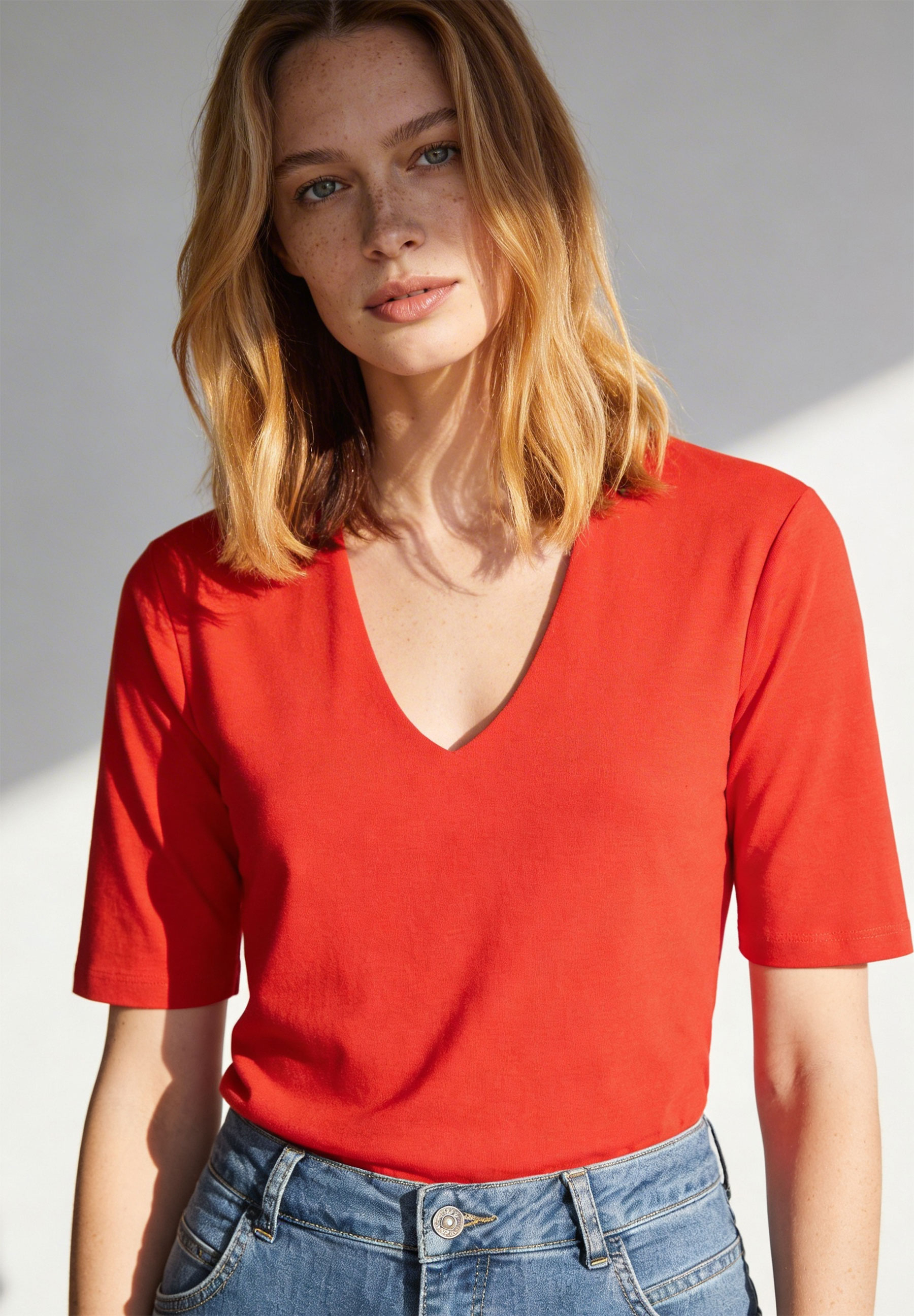 Eine Frau trägt ein rotes MORE & MORE T-Shirt mit V-Ausschnitt und Jeans. Das Shirt hat eine Jersey-Materialkonstruktion.