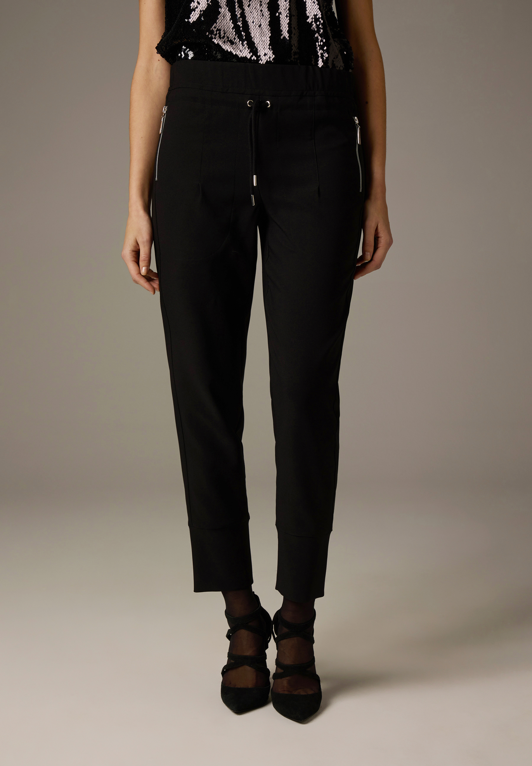Eine Person trägt schwarze MORE & MORE Hose mit Reißverschlüssen und dazu passende High Heels.