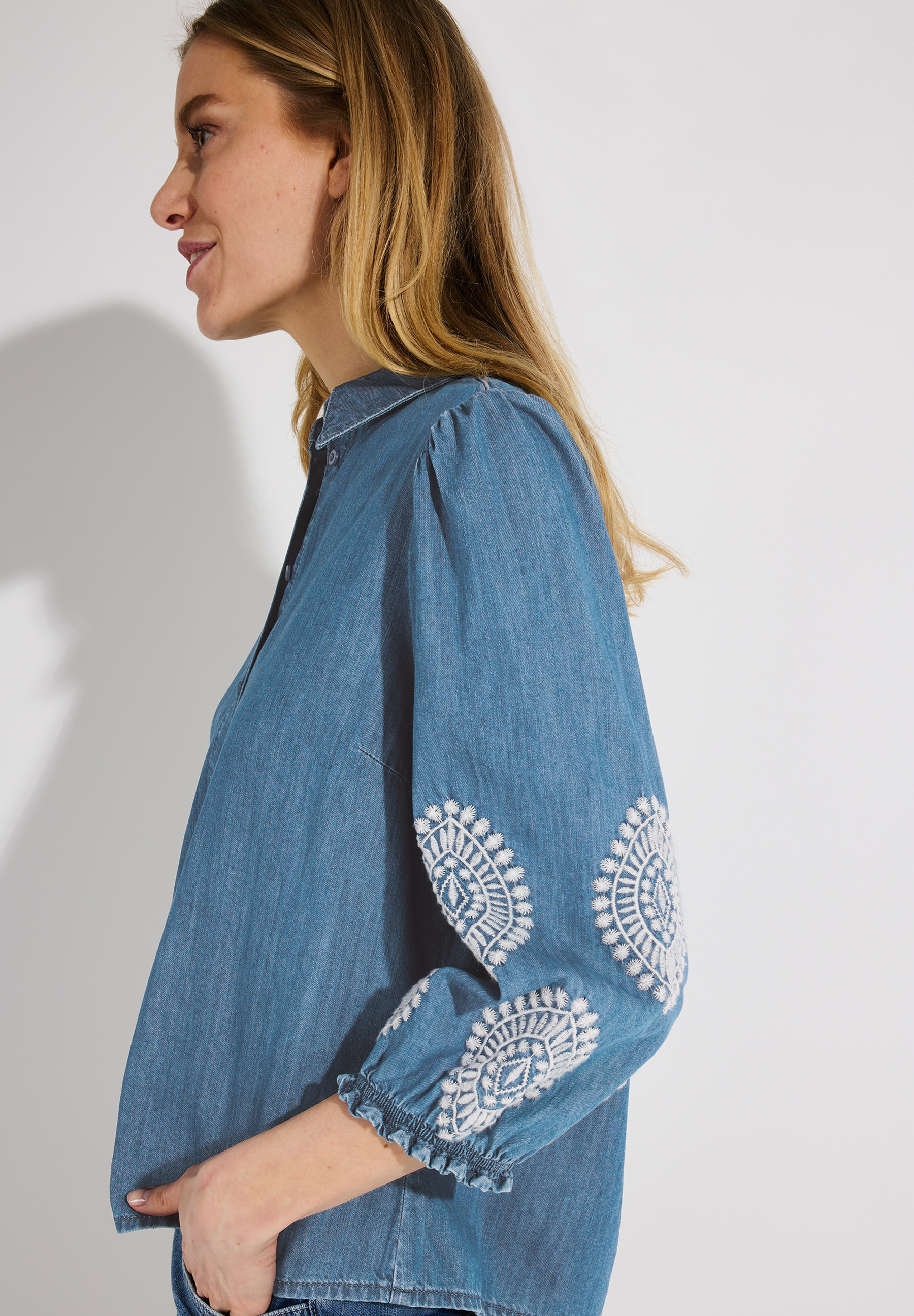 Eine blaue Denim-Bluse von MORE & MORE mit weißer Stickerei an den Ärmeln, seitliche Profilansicht. Eine blaue Denim-Bluse von MORE & MORE mit weißer Stickerei an den Ärmeln, seitliche Profilansicht.