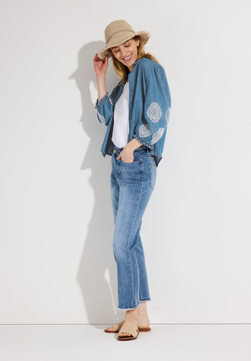 Eine lächelnde Frau mit Hut, Jeans und MORE & MORE Jeansbluse mit weißer Stickerei. Eine lächelnde Frau mit Hut, Jeans und MORE & MORE Jeansbluse mit weißer Stickerei.