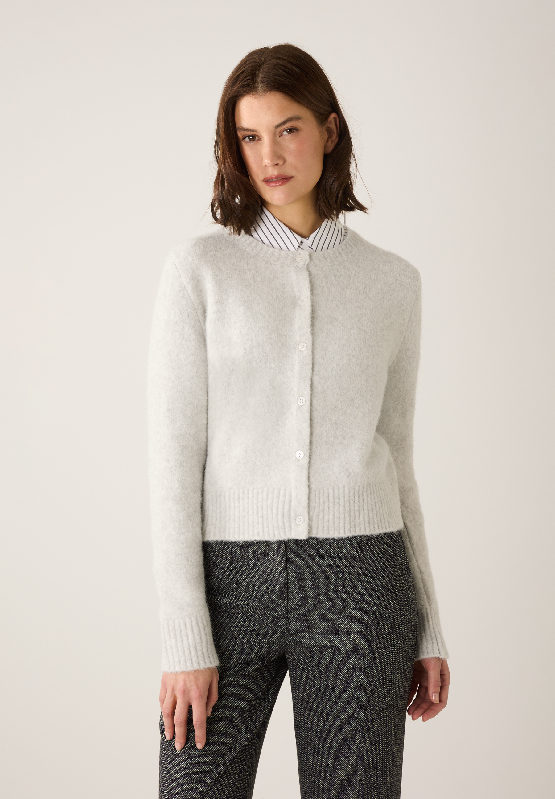 Eine Frau mit einem MORE & MORE Oberteil: Strickjacke in Grau über einer gestreiften Bluse und einer grauen Hose.