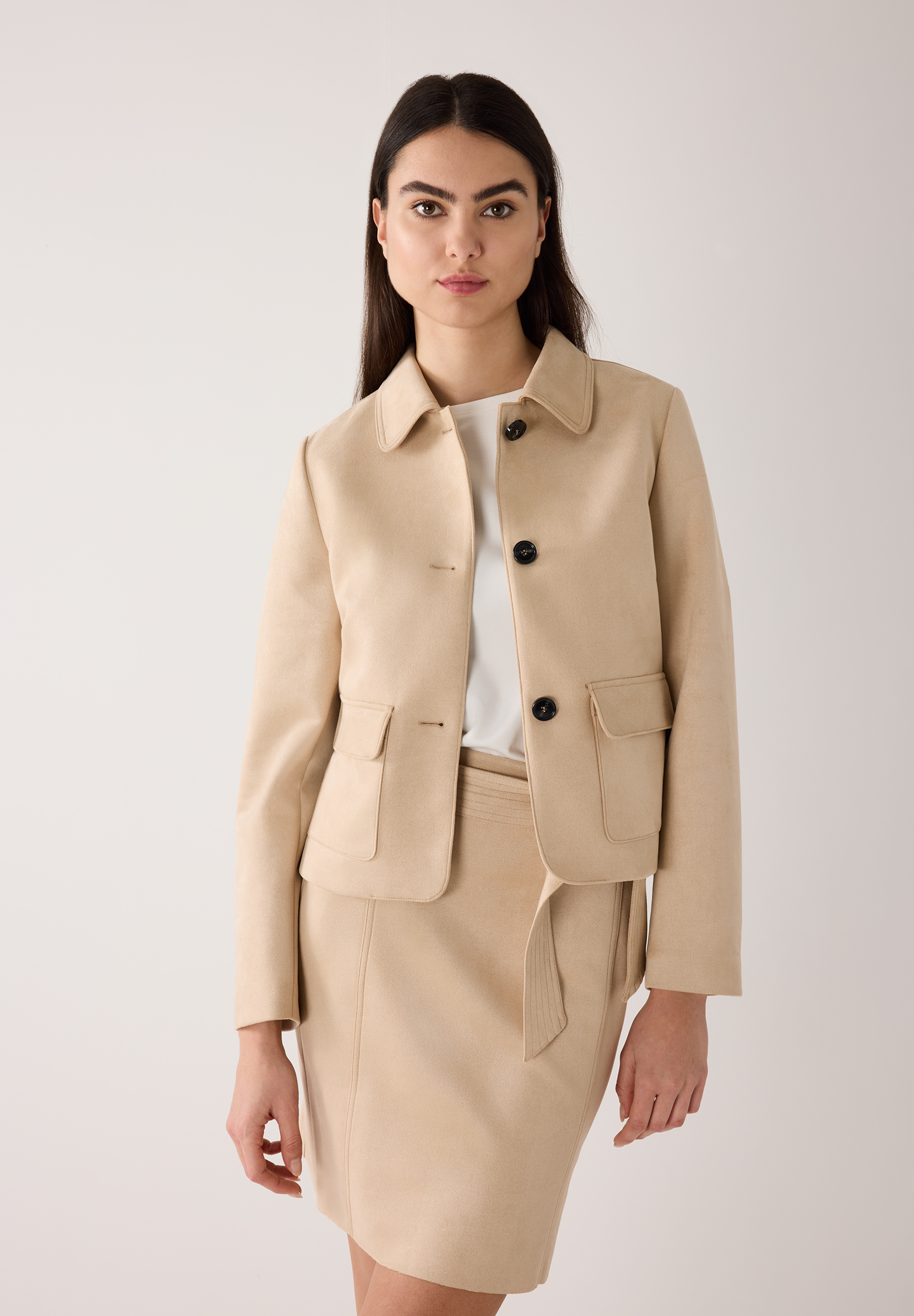 Eine Frau mit MORE & MORE Blazer aus Kunstleder und passendem Rock vor hellem Hintergrund.