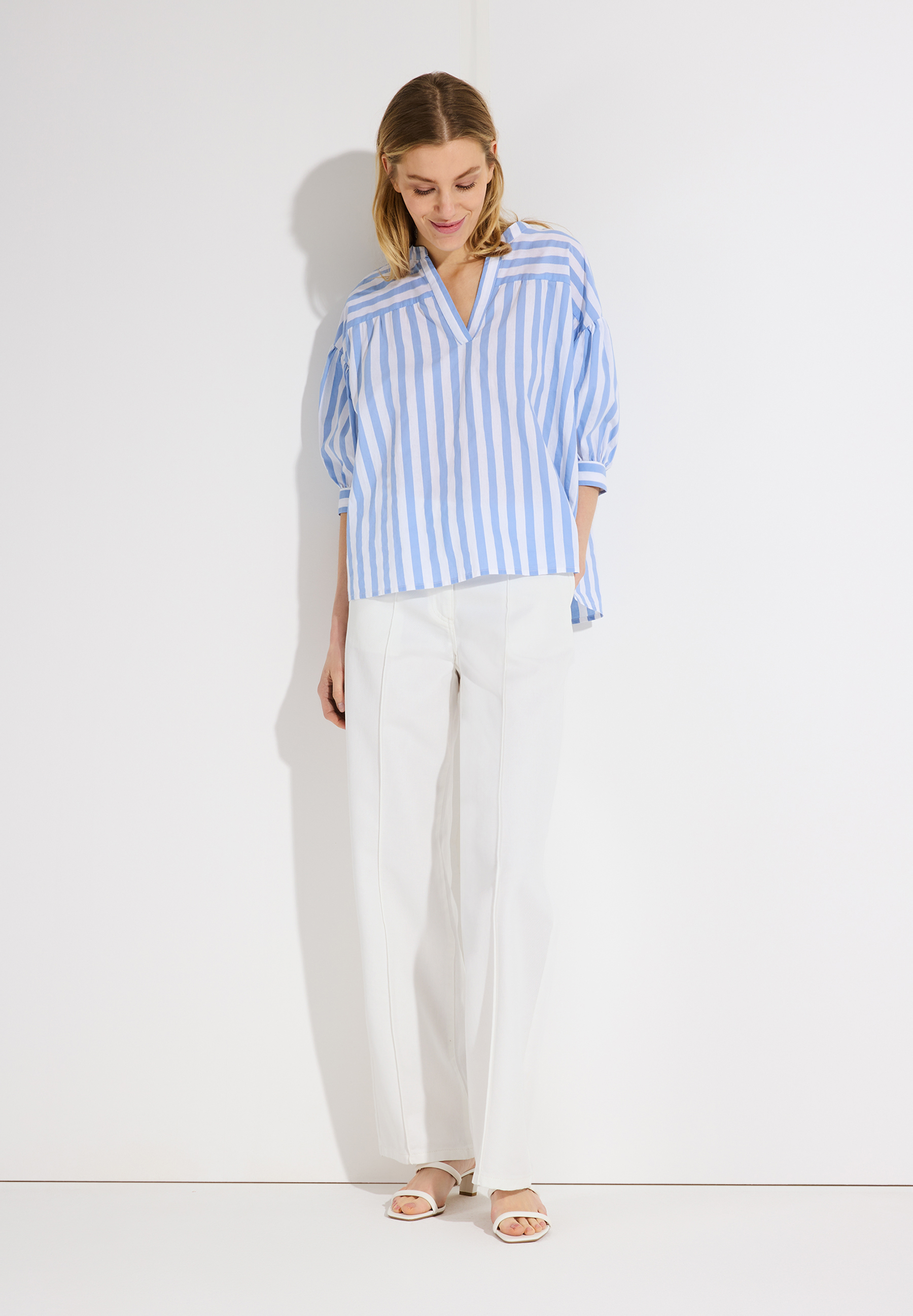 Eine Frau in MORE & MORE-Popeline-Bluse mit blauen Streifen und weißer Hose vor einem weißen Hintergrund. Eine Frau in MORE & MORE-Popeline-Bluse mit blauen Streifen und weißer Hose vor einem weißen Hintergrund.