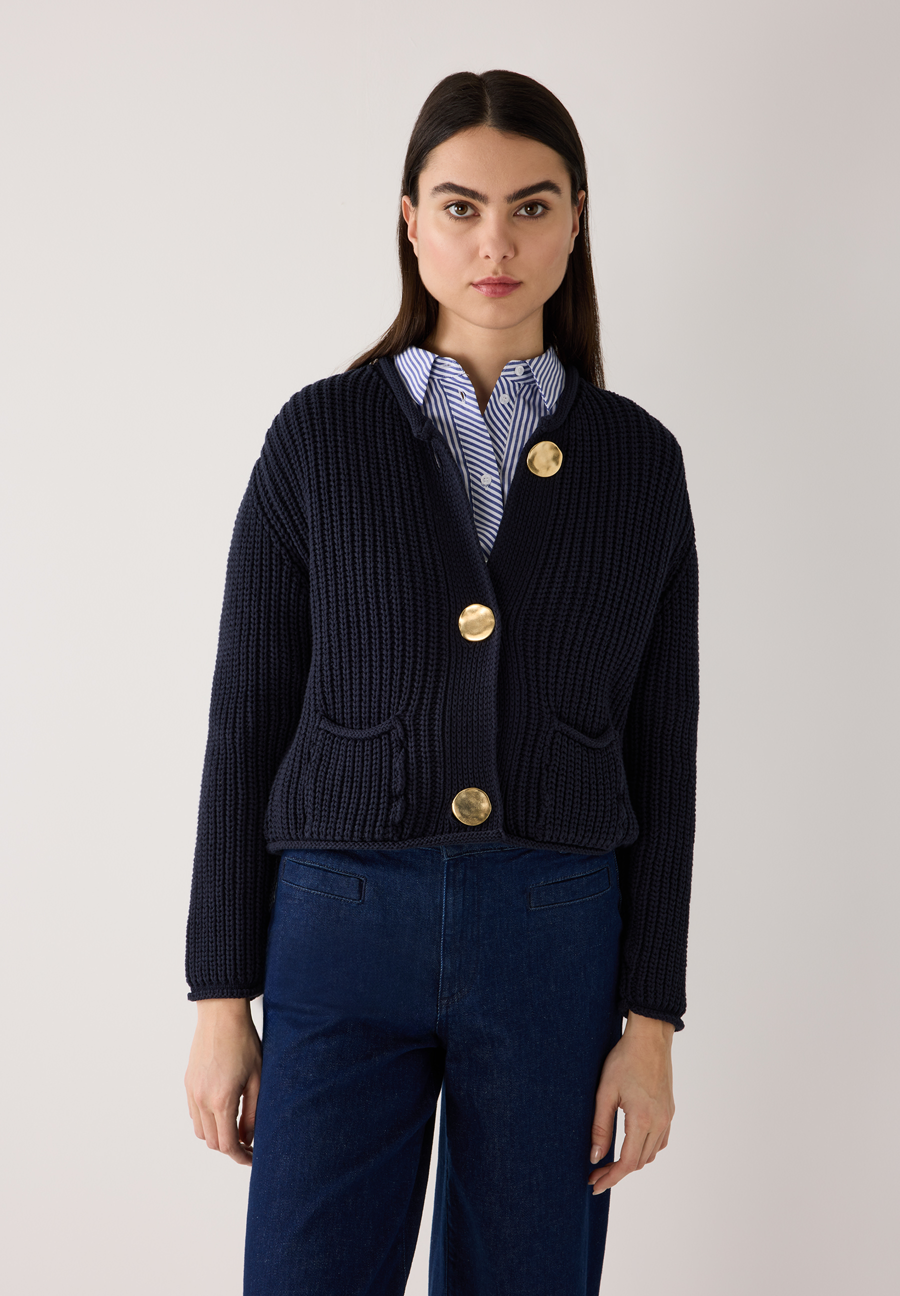 Eine Frau in einem MORE & MORE Rippstrick-Cardigan mit goldenen Knöpfen und blauer Hose. Eine Frau in einem MORE & MORE Rippstrick-Cardigan mit goldenen Knöpfen und blauer Hose.