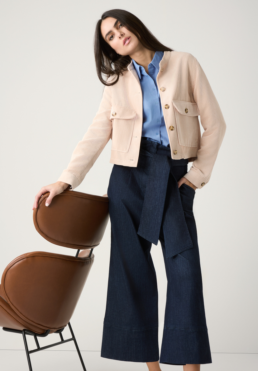 Eine Frau in MORE & MORE Oberteilen, Popeline-Bluse und Cordjacke, posiert an einem Stuhl. Eine Frau in MORE & MORE Oberteilen, Popeline-Bluse und Cordjacke, posiert an einem Stuhl.