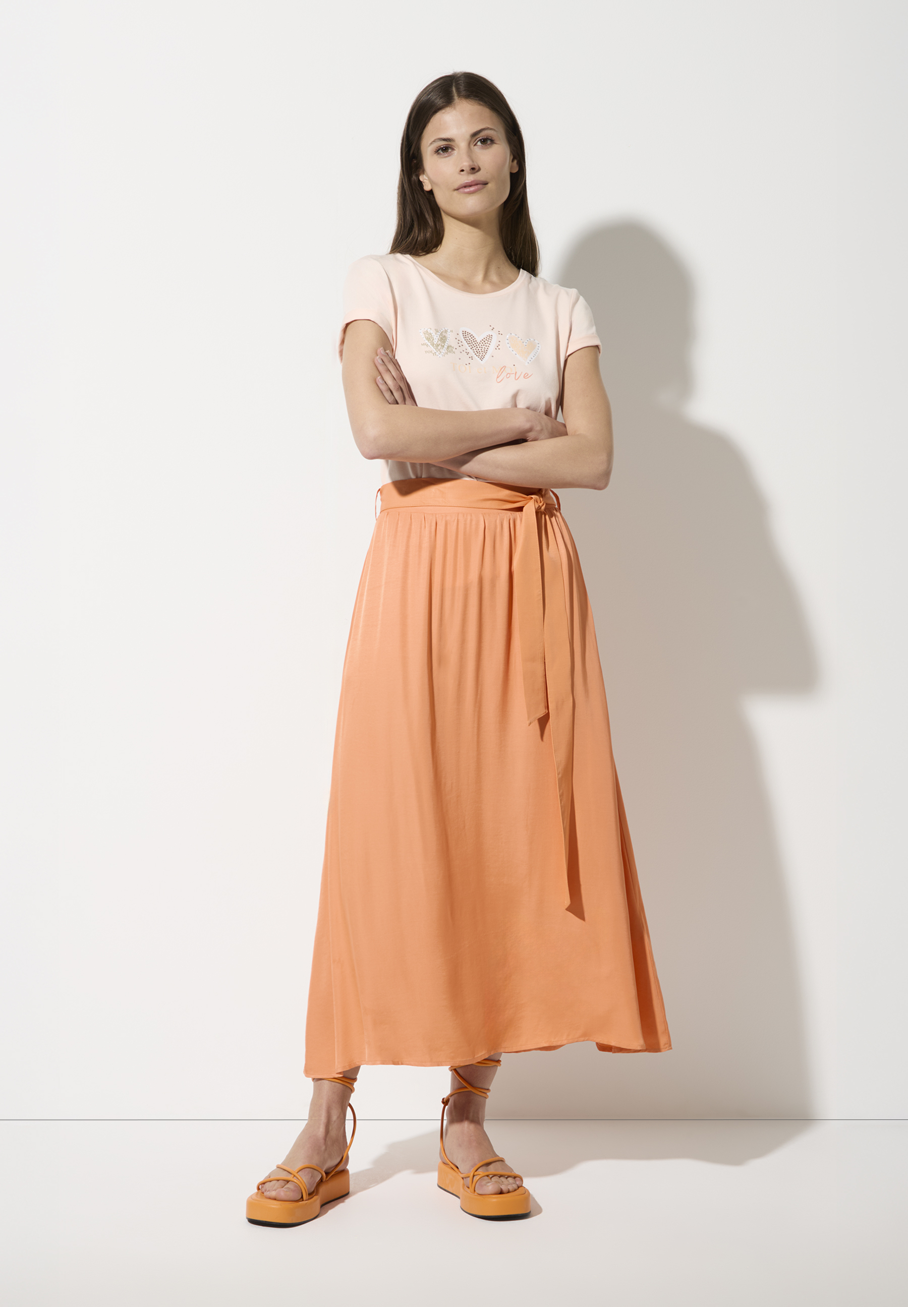 Eine Frau, die einen orangefarbenen Rock von MORE & MORE trägt, steht vor einer weißen Wand. Eine Frau, die einen orangefarbenen Rock von MORE & MORE trägt, steht vor einer weißen Wand.