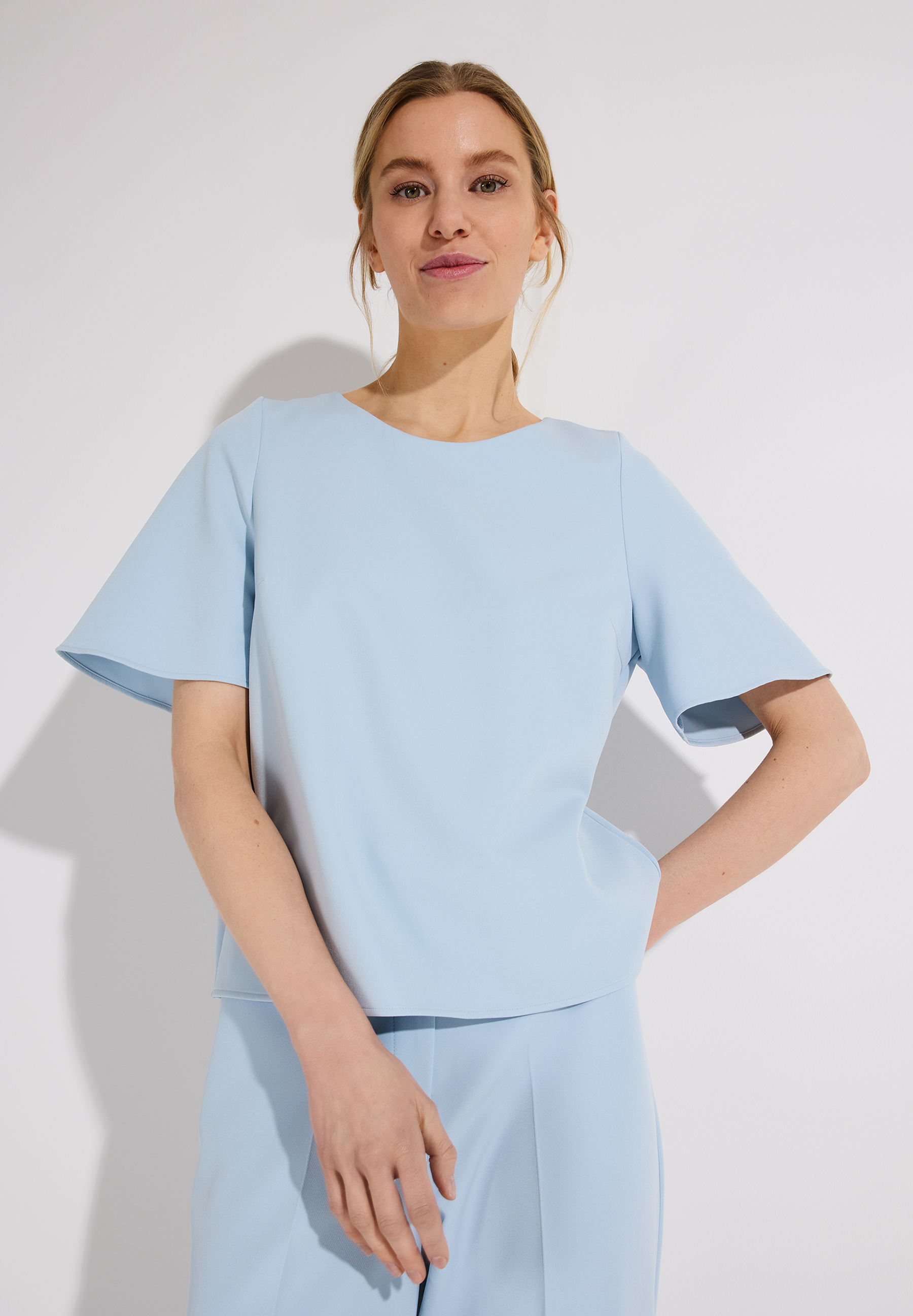 Eine Frau trägt ein hellblaues Oberteil von MORE & MORE. Bluse mit kurzen, ausgestellten Ärmeln.