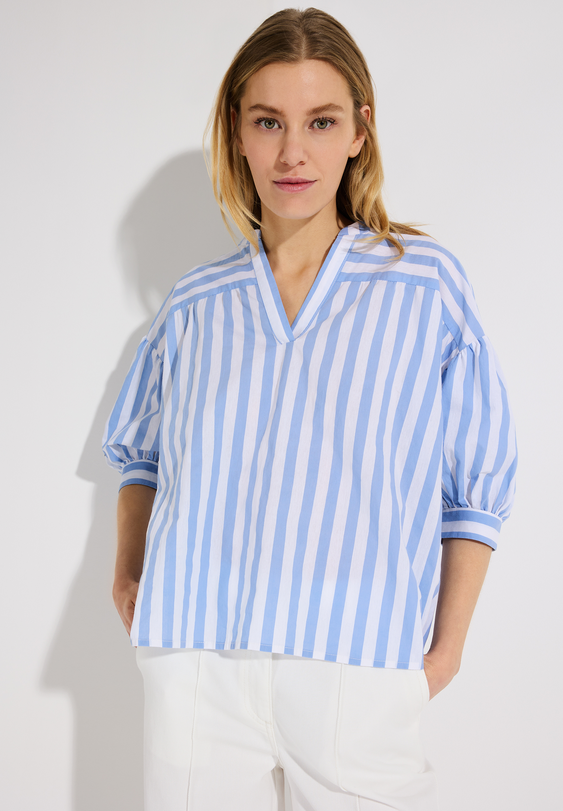 Eine Frau trägt eine MORE & MORE Bluse mit blau-weißen Streifen und eine weiße Hose.