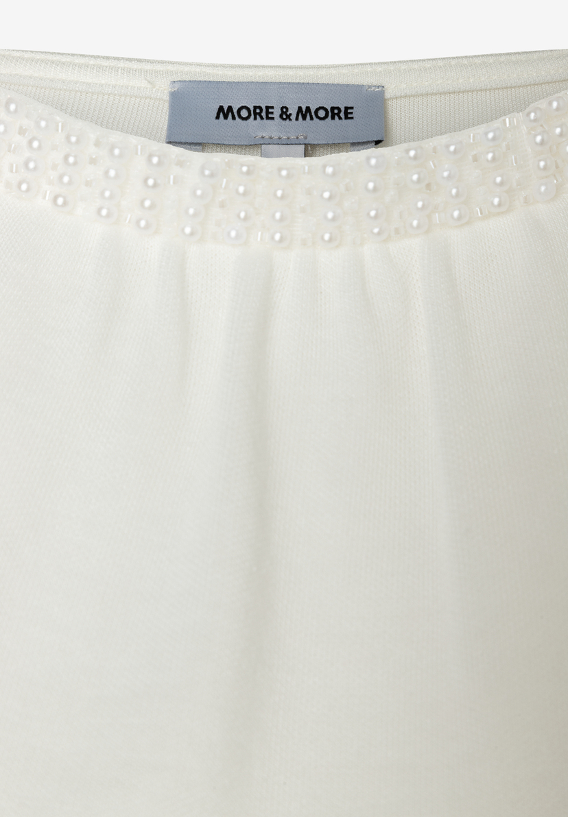 Detailansicht eines MORE & MORE Oberteils mit Jersey-Material und Perlenbesatz am Kragen. Detailansicht eines MORE & MORE Oberteils mit Jersey-Material und Perlenbesatz am Kragen.