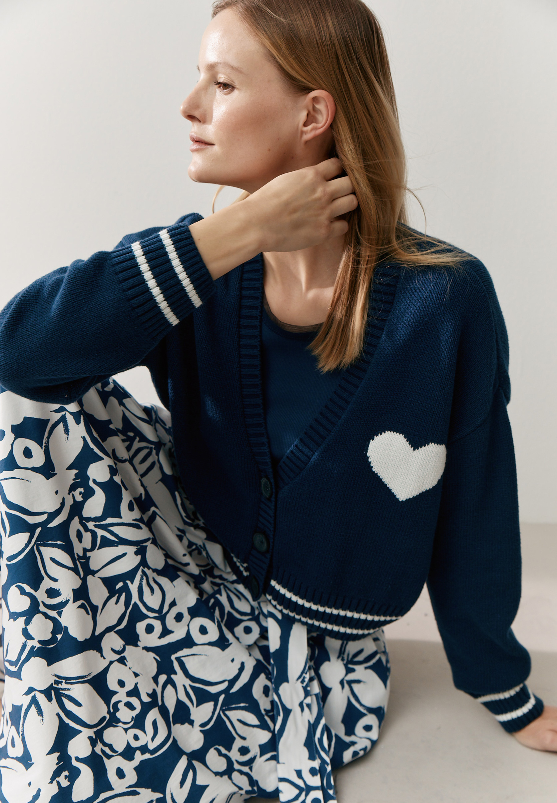 Eine Frau trägt einen marineblauen MORE & MORE Strick-Cardigan mit weißem Herz und Blumenhose. Eine Frau trägt einen marineblauen MORE & MORE Strick-Cardigan mit weißem Herz und Blumenhose.