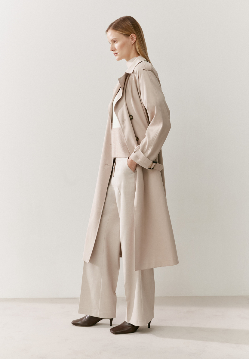 Eine Frau trägt einen hellen MORE & MORE Trenchcoat und eine passende Hose vor einem weißen Hintergrund. Eine Frau trägt einen hellen MORE & MORE Trenchcoat und eine passende Hose vor einem weißen Hintergrund.
