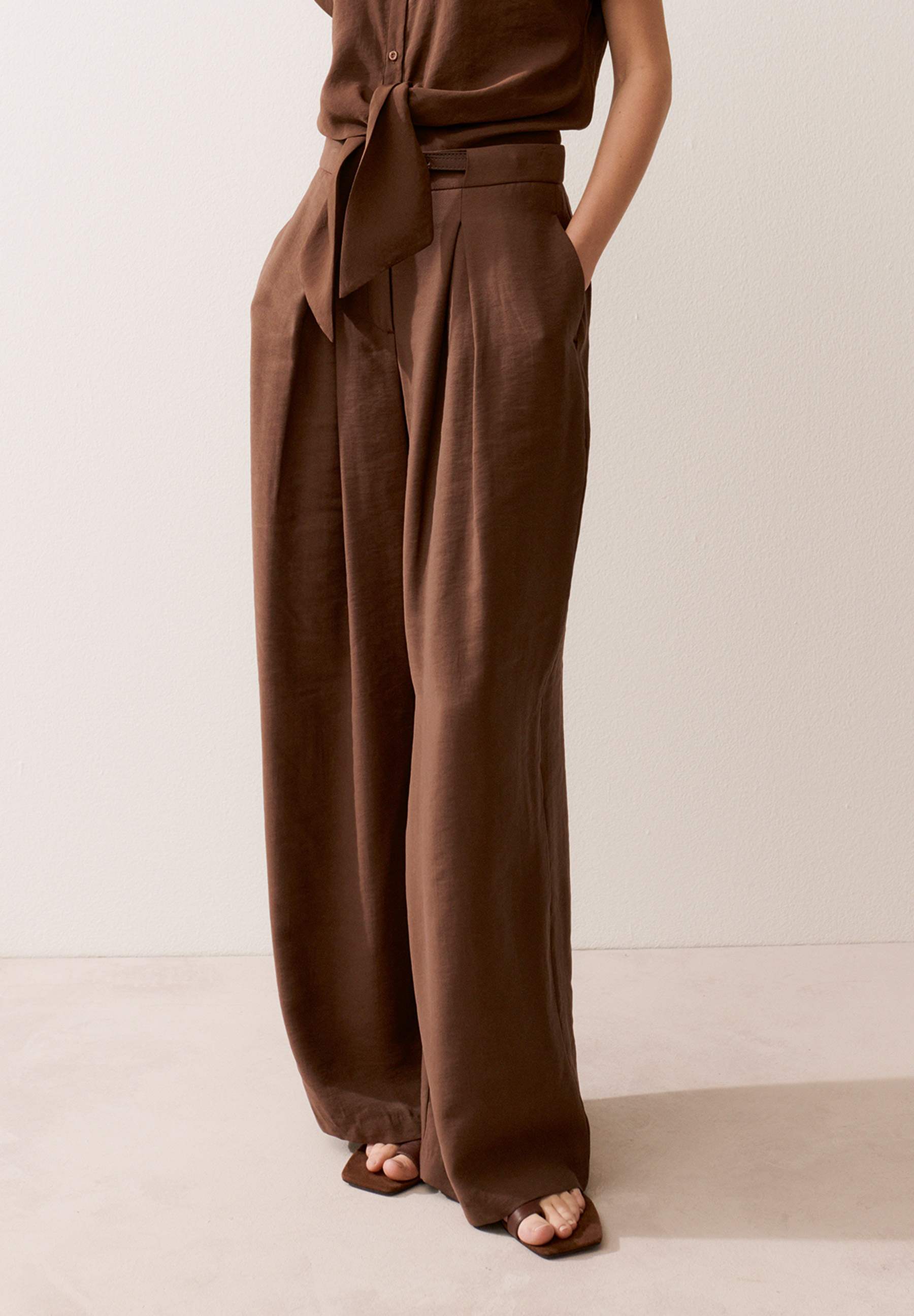 Eine Frau trägt MORE & MORE-Hosen. Die Hose ist braun und hat weite Beine. Ihre Hände stecken in den Hosentaschen.