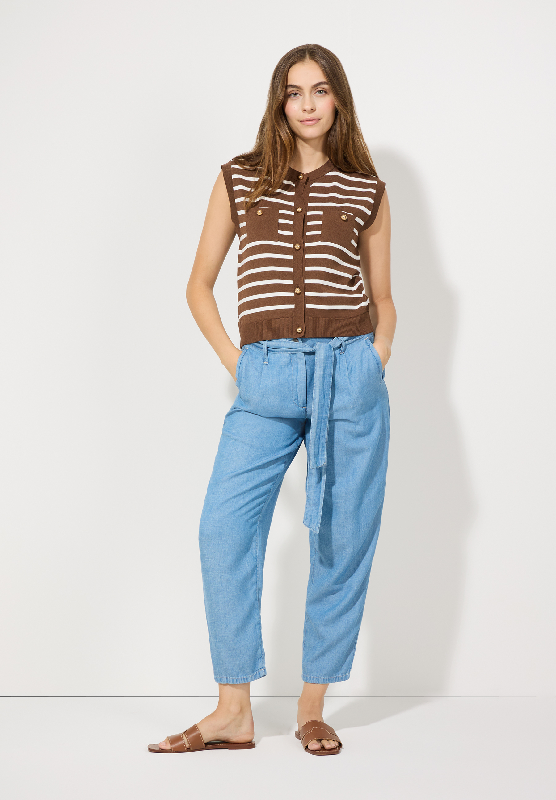 Eine Frau trägt MORE & MORE Denim-Hosen mit Bindegürtel und ein braun-weiß gestreiftes Top. Eine Frau trägt MORE & MORE Denim-Hosen mit Bindegürtel und ein braun-weiß gestreiftes Top.