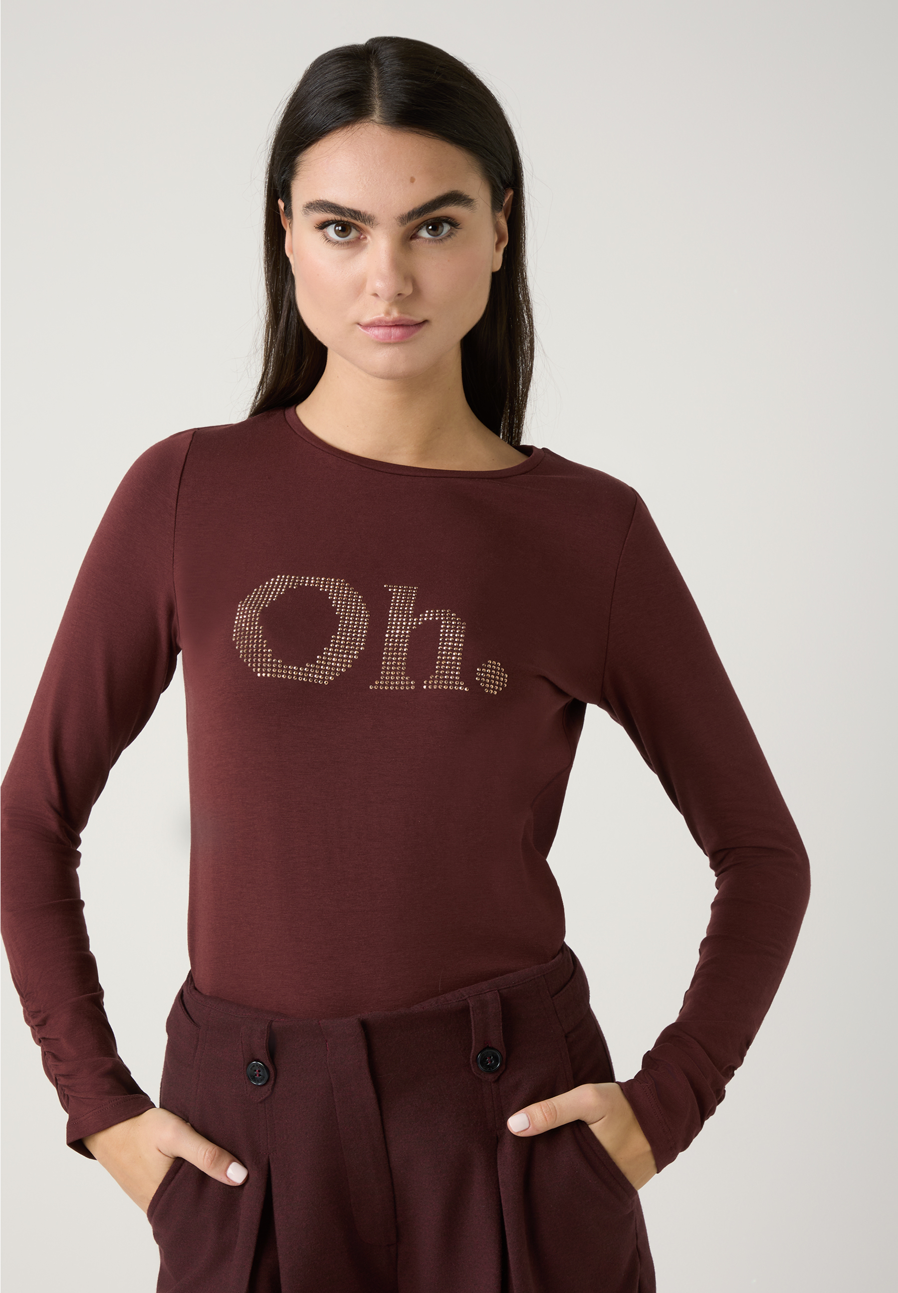 Eine Frau mit einem bordeauxroten, langärmeligen MORE & MORE T-Shirt mit dem glitzernden Schriftzug "Oh." posiert vor hellem Hintergrund.