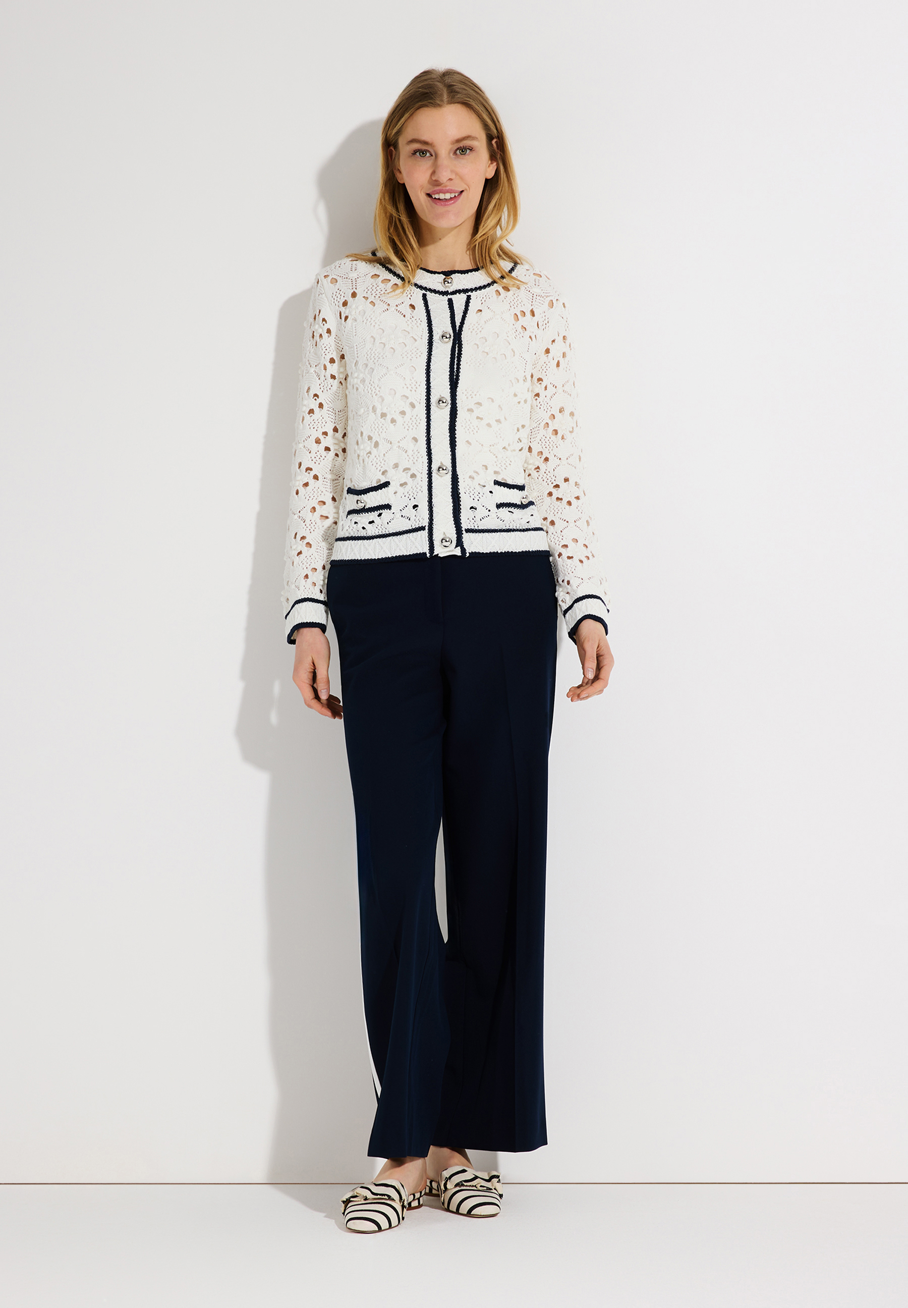 Eine blonde Frau, die einen transparenten Lochstrick-Cardigan von MORE & MORE und eine marineblaue Hose vor einem weißen Hintergrund trägt. Eine blonde Frau, die einen transparenten Lochstrick-Cardigan von MORE & MORE und eine marineblaue Hose vor einem weißen Hintergrund trägt.