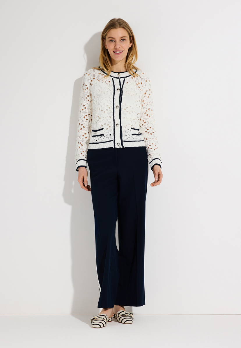 Eine blonde Frau, die einen transparenten Lochstrick-Cardigan von MORE & MORE und eine marineblaue Hose vor einem weißen Hintergrund trägt. Eine blonde Frau, die einen transparenten Lochstrick-Cardigan von MORE & MORE und eine marineblaue Hose vor einem weißen Hintergrund trägt.