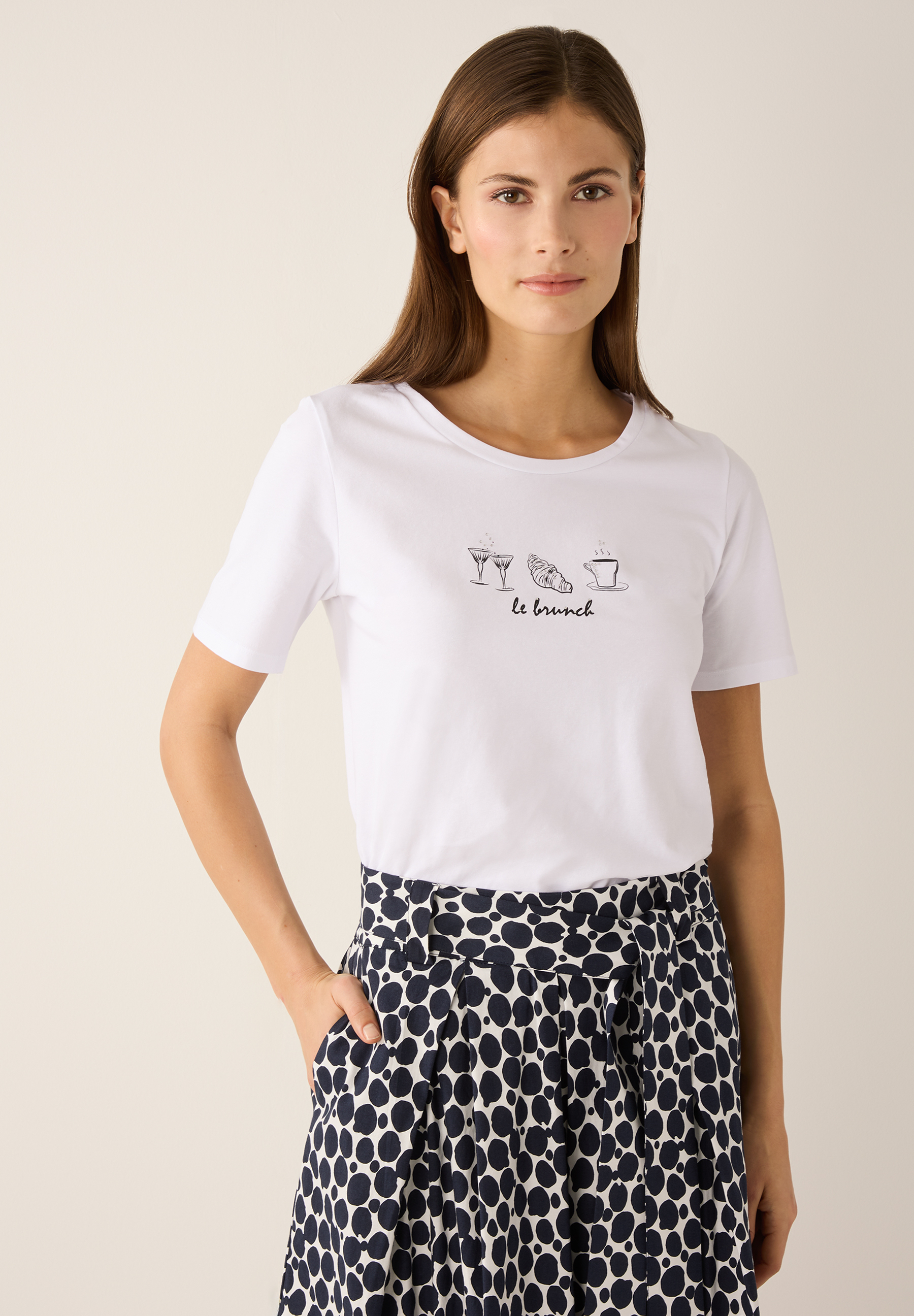 Eine Frau trägt ein weißes MORE & MORE T-Shirt mit "Le Brunch"-Motiv und einen Rock mit großen Punkten.