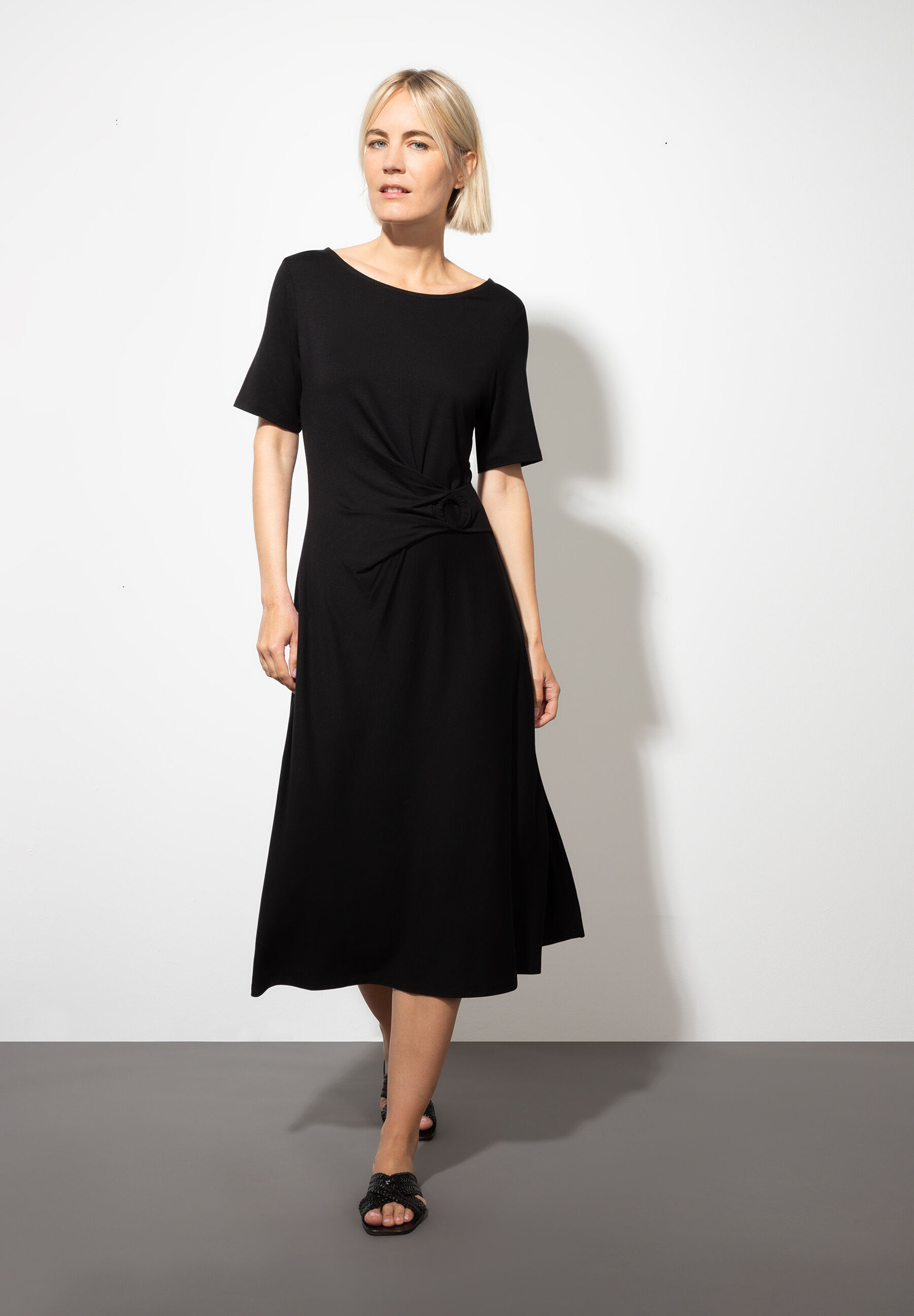 Eine Frau in einem schwarzen MORE & MORE Jersey-Kleid mit komfortablem, elastischem Stoff posiert vor einer weißen Wand.