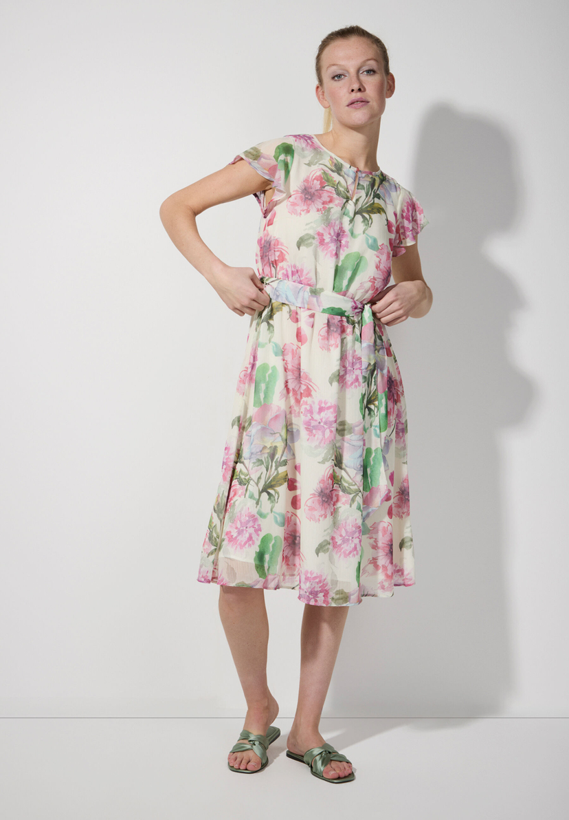 Eine Frau in einem MORE & MORE Chiffon-Kleid mit Blumenmuster posiert vor einer weißen Wand. Eine Frau in einem MORE & MORE Chiffon-Kleid mit Blumenmuster posiert vor einer weißen Wand.