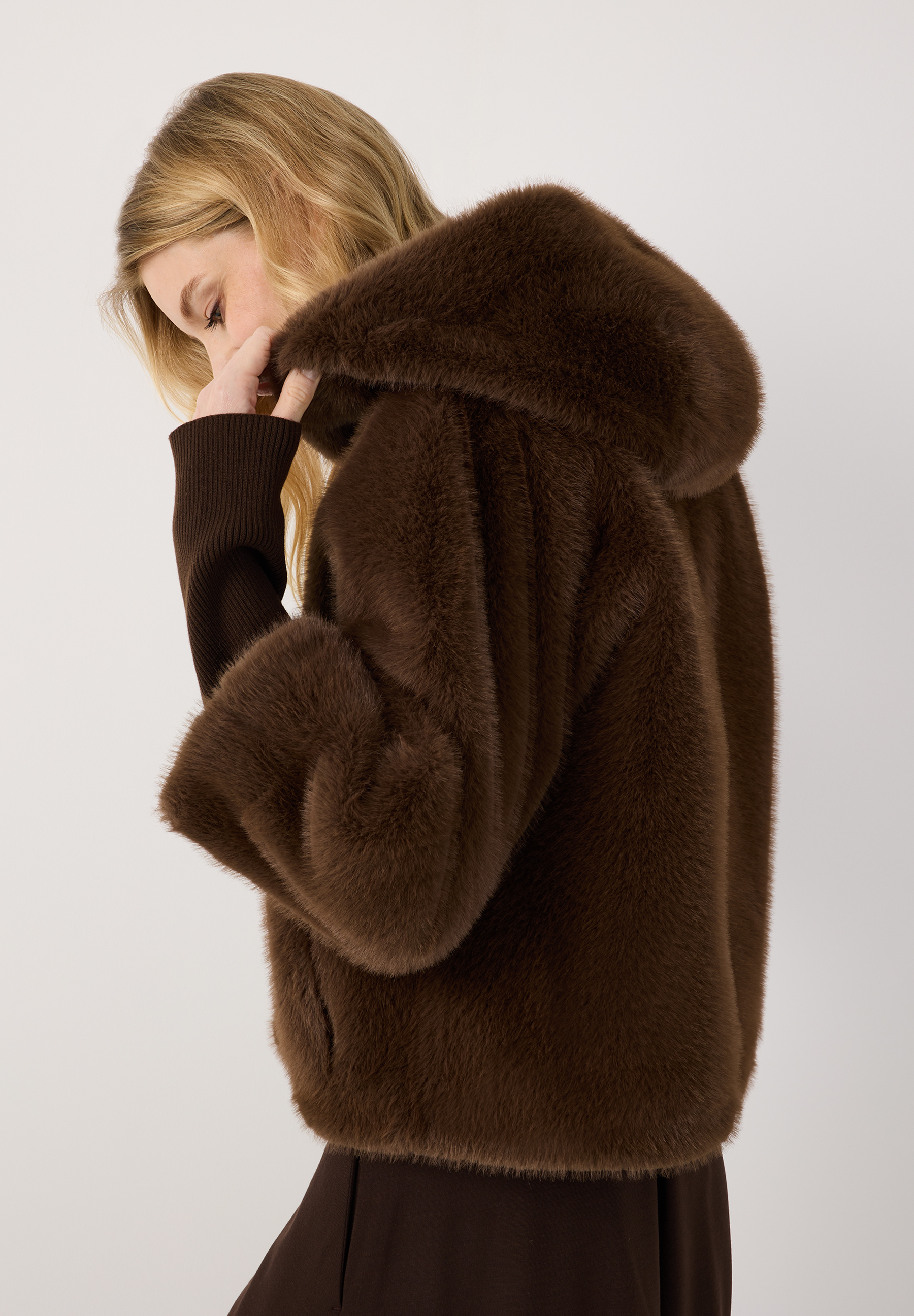 MORE & MORE Jacke aus braunem Kunstpelz mit Kapuze, gestrickten Ärmelbündchen und einer blonden Frau in Profilansicht.