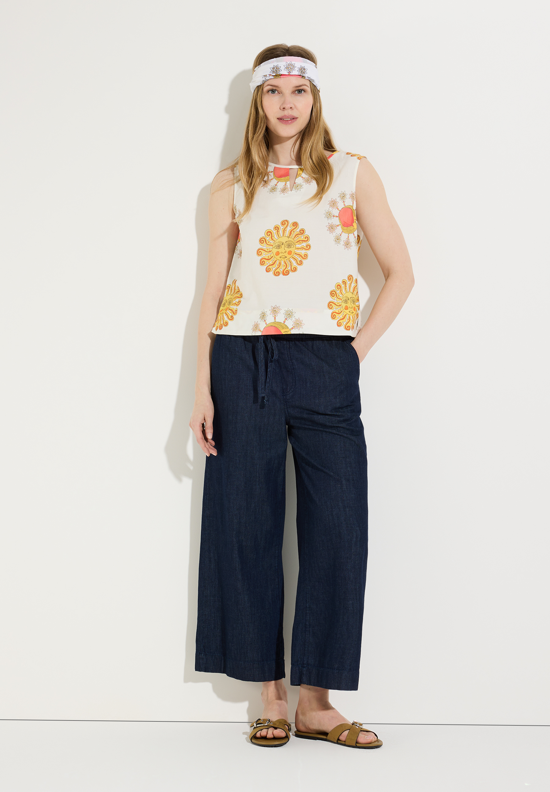 Eine Frau mit Stirnband und MORE & MORE Bluse mit Sonnenmuster posiert vor einer weißen Wand.