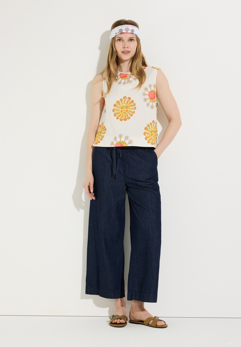 Eine Frau mit Stirnband und MORE & MORE Bluse mit Sonnenmuster posiert vor einer weißen Wand.