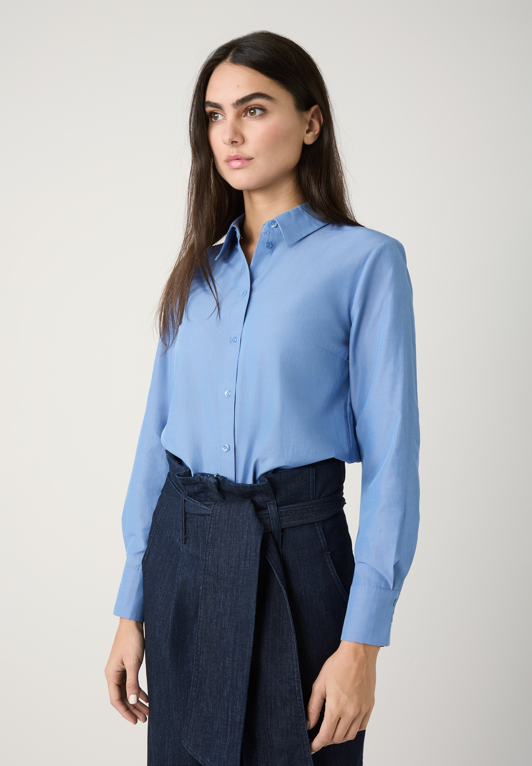 Eine Frau trägt eine blaue Popeline-Bluse von MORE & MORE und einen Jeansrock mit Bindegürtel.