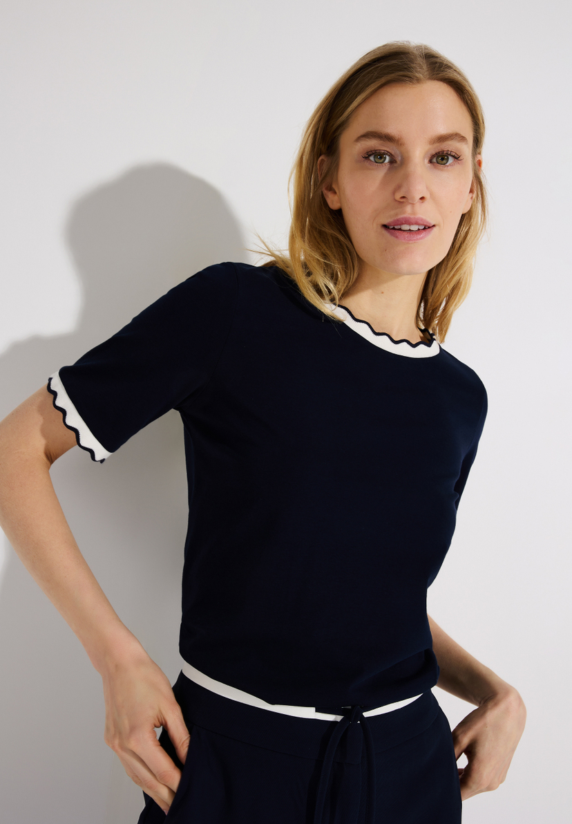 Eine Frau trägt ein dunkelblaues MORE & MORE T-Shirt mit weißen Rüschen am Hals und an den Ärmeln vor einem weißen Hintergrund. Eine Frau trägt ein dunkelblaues MORE & MORE T-Shirt mit weißen Rüschen am Hals und an den Ärmeln vor einem weißen Hintergrund.