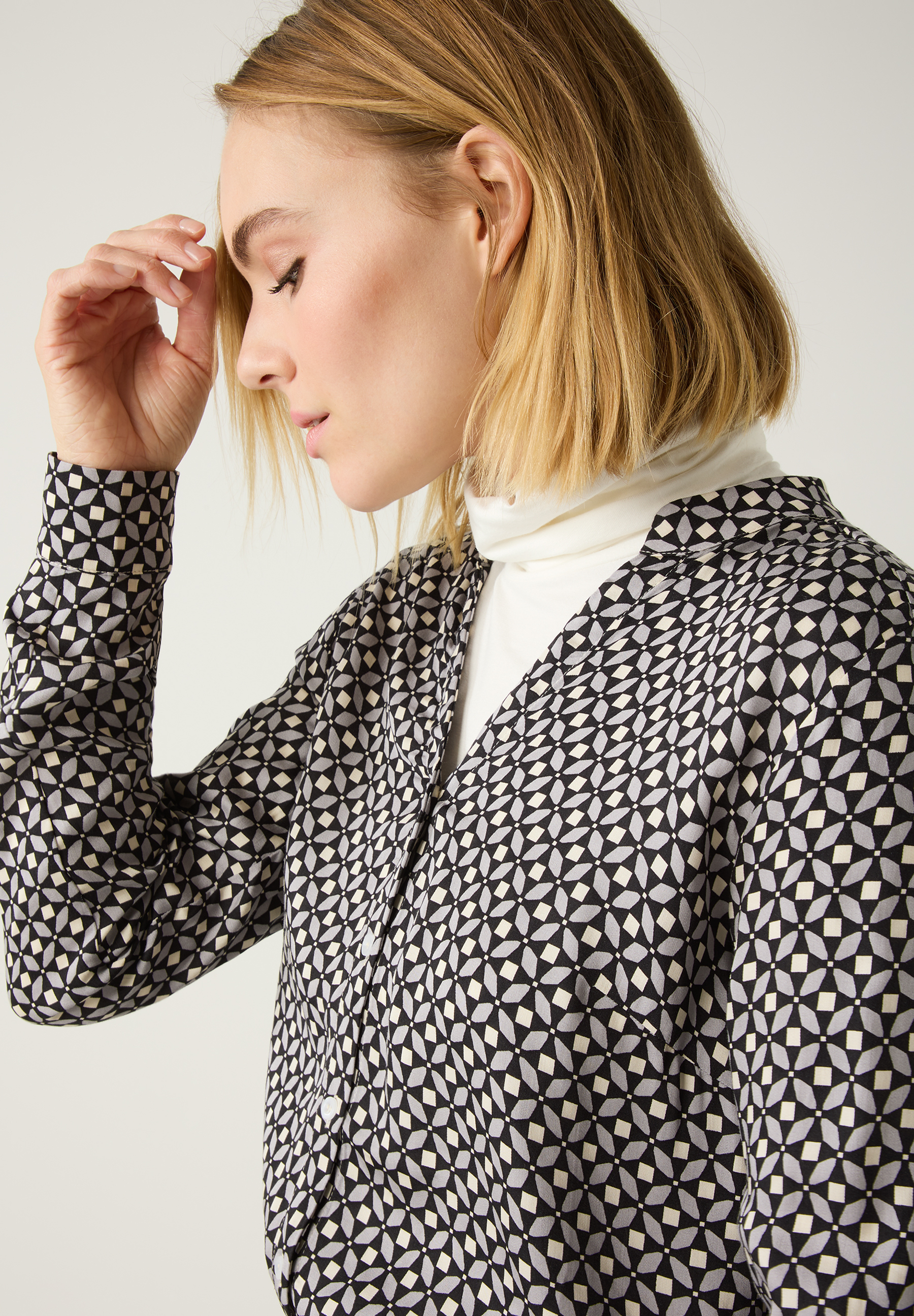 Eine Frau mit einer MORE & MORE Bluse mit geometrischem Muster und Rollkragenpullover darunter. Eine Frau mit einer MORE & MORE Bluse mit geometrischem Muster und Rollkragenpullover darunter.