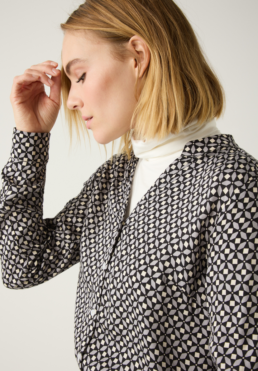 Eine Frau mit einer MORE & MORE Bluse mit geometrischem Muster und Rollkragenpullover darunter. Eine Frau mit einer MORE & MORE Bluse mit geometrischem Muster und Rollkragenpullover darunter.
