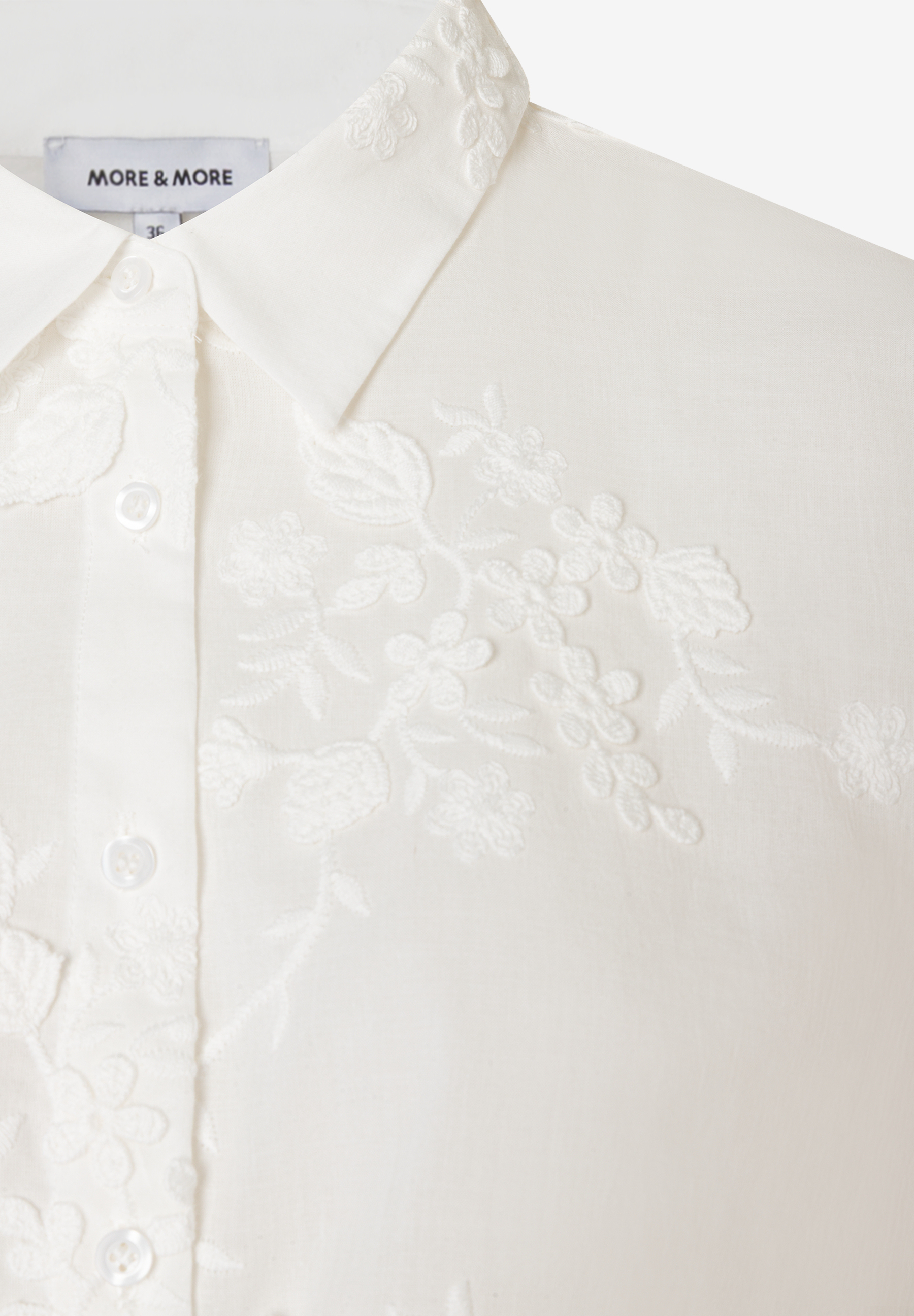 Detailaufnahme einer weißen MORE & MORE Bluse mit filigraner Blumenstickerei und Knopfleiste.