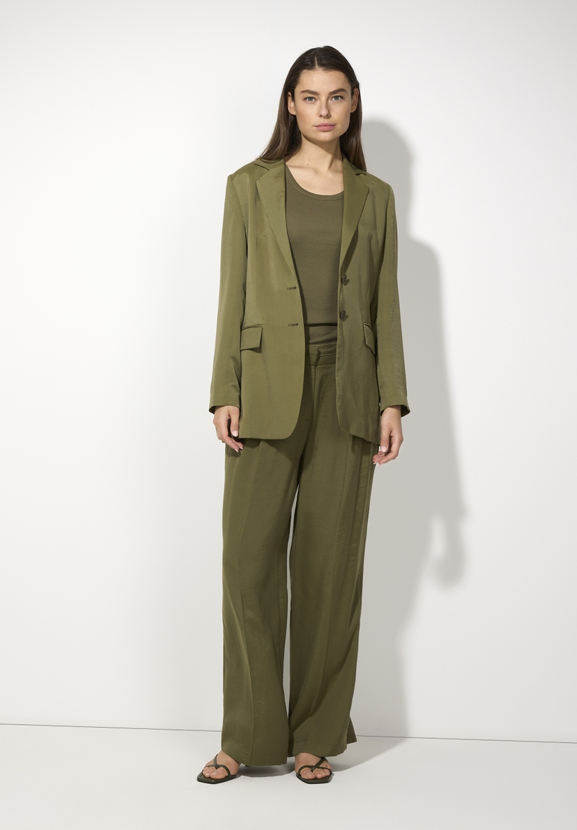 Eine Frau trägt einen olivgrünen Blazer von MORE & MORE mit passender Hose und einem T-Shirt vor einem weißen Hintergrund. Eine Frau trägt einen olivgrünen Blazer von MORE & MORE mit passender Hose und einem T-Shirt vor einem weißen Hintergrund.