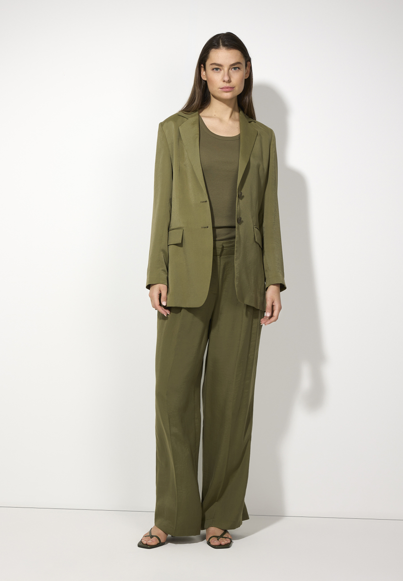 Eine Frau trägt einen olivgrünen Blazer von MORE & MORE mit passender Hose und einem T-Shirt vor einem weißen Hintergrund. Eine Frau trägt einen olivgrünen Blazer von MORE & MORE mit passender Hose und einem T-Shirt vor einem weißen Hintergrund.