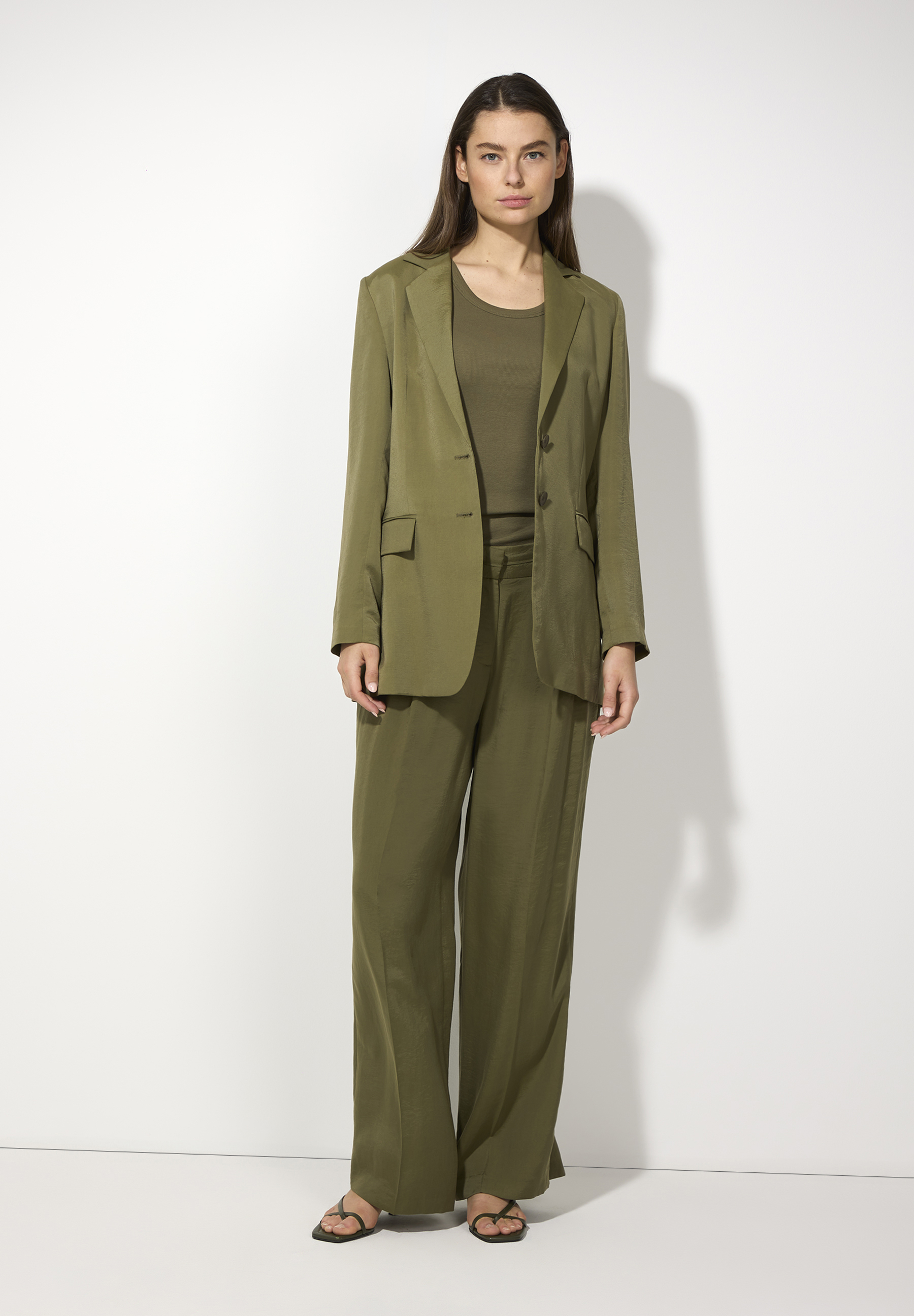 Eine Frau trägt einen olivgrünen Blazer von MORE & MORE mit passender Hose und einem T-Shirt vor einem weißen Hintergrund. Eine Frau trägt einen olivgrünen Blazer von MORE & MORE mit passender Hose und einem T-Shirt vor einem weißen Hintergrund.