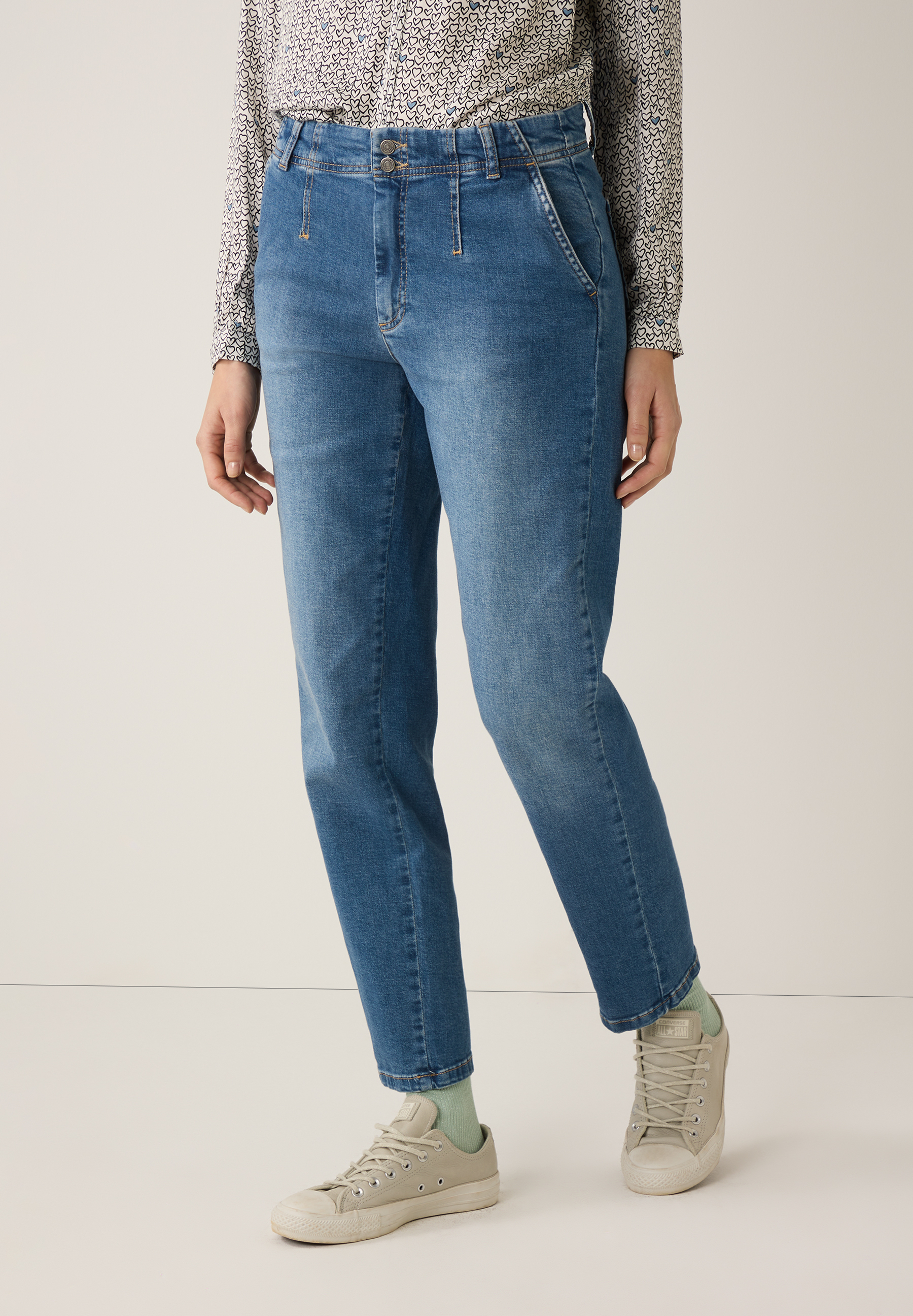 Eine Frau trägt MORE & MORE Jeans, kombiniert mit einem gemusterten Hemd und beigen Turnschuhen vor einem neutralen Hintergrund.