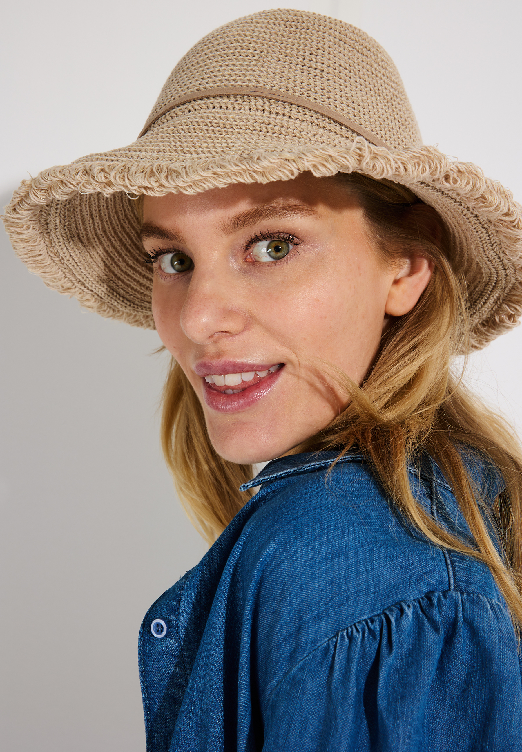 Eine Frau mit einem beigen MORE & MORE Hut lächelt in die Kamera.