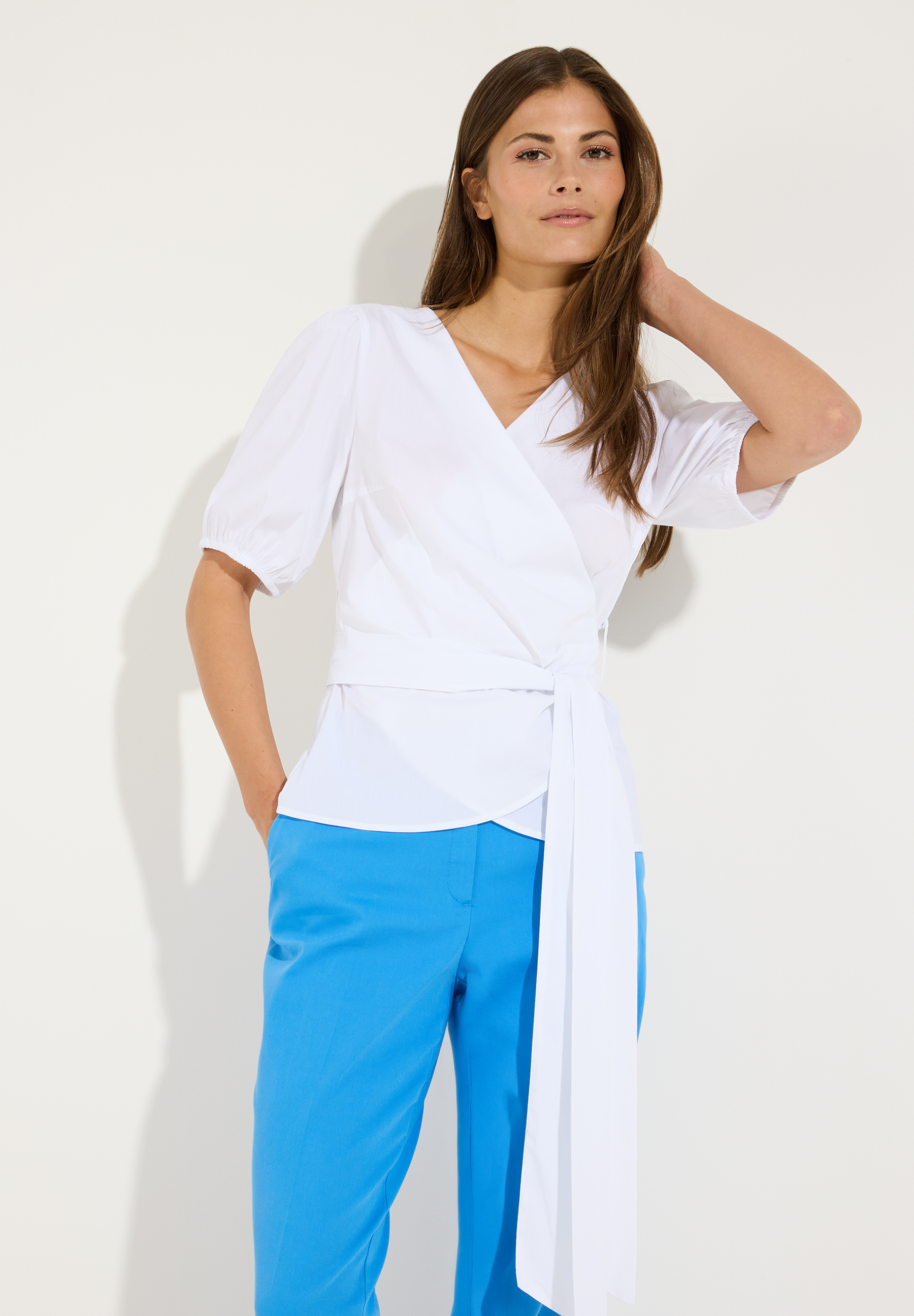 MORE & MORE Wickelbluse in Weiß mit V-Ausschnitt, 3/4-Ärmeln und Taillenband, kombiniert mit blauer Hose. MORE & MORE Wickelbluse in Weiß mit V-Ausschnitt, 3/4-Ärmeln und Taillenband, kombiniert mit blauer Hose.