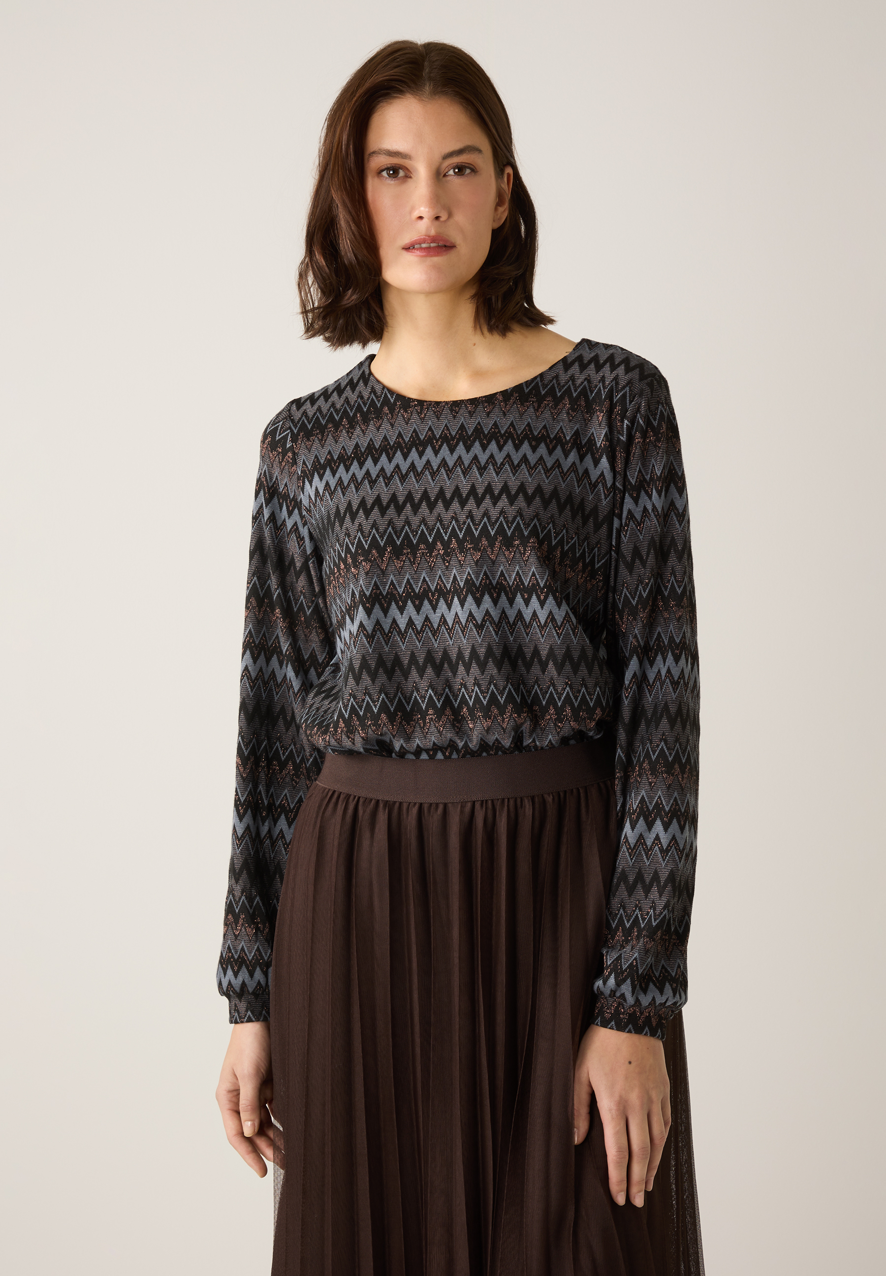 Eine Frau, die ein MORE & MORE-Oberteil mit Zickzackmuster und einen braunen Faltenrock trägt.