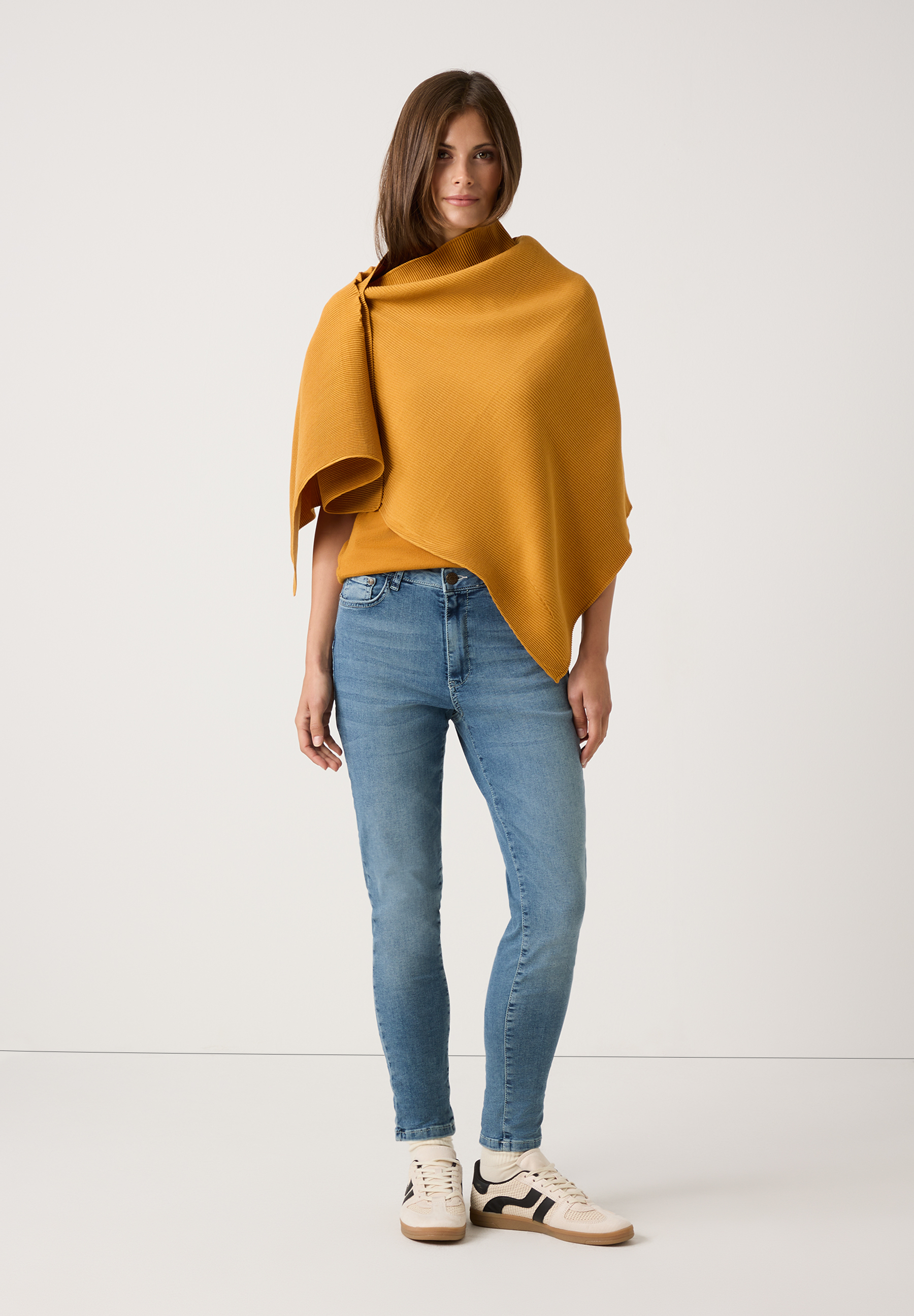 Eine Frau in MORE & MORE Accessoires: ockergelbes Tuch, Jeans und Turnschuhe vor weißem Hintergrund.