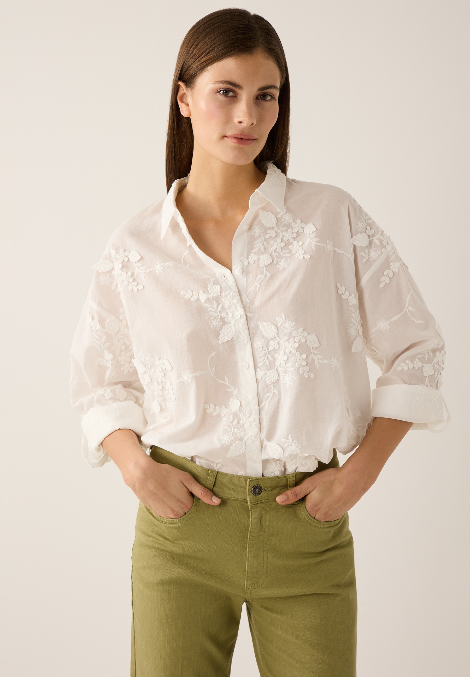 Eine Frau mit einer weißen MORE & MORE Bluse mit Blumenmuster und grüner Hose. Eine Frau mit einer weißen MORE & MORE Bluse mit Blumenmuster und grüner Hose.